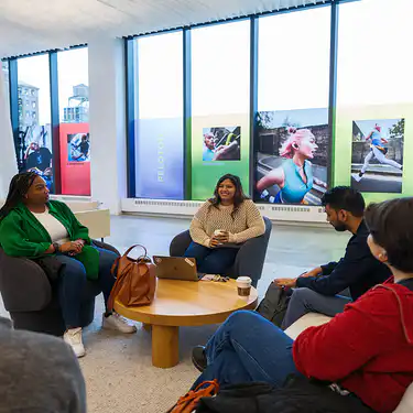 Five Peloton team members sitting on chairs in a common office workspace engage in conversation.