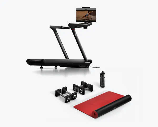 Peloton Tread treadmill displayed alongside two pairs of dumbbells, a water bottle, and a rolled-out exercise mat.