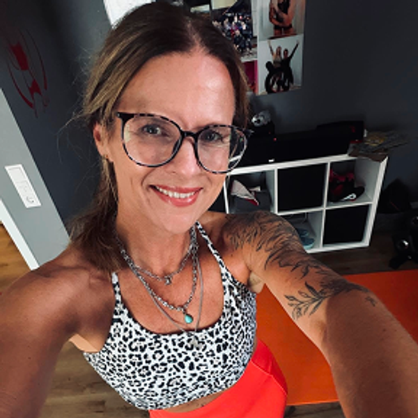 Woman with glasses and tattoos smiling while taking a selfie in a gym setting.