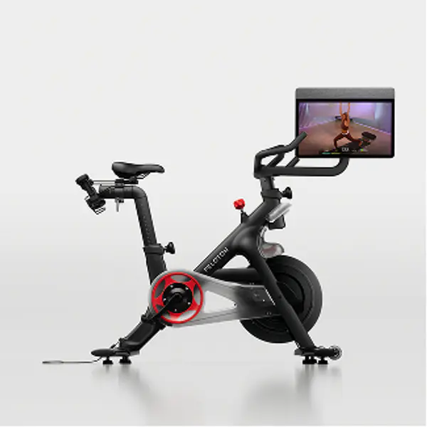 Side view of a Peloton Bike showing the seat, handlebars, flywheel, and attached screen displaying a workout class.