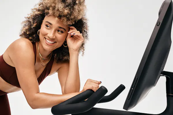 Cycling instructor smiling and leaning against the screen of a Peloton exercise bike.
