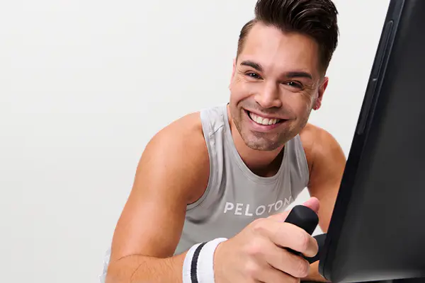 Cycling instructor smiling and leaning against the screen of a Peloton exercise bike.