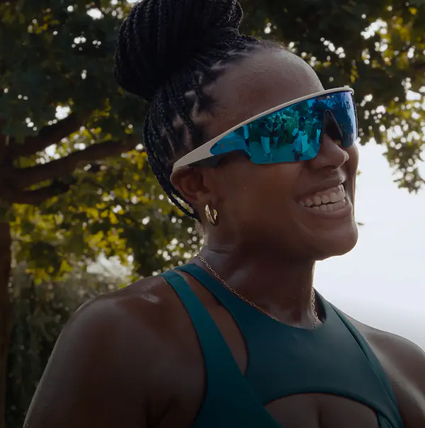 Close-up portrait of Raquel smiling outdoors, wearing sports sunglasses and a green tank top with her hair in a bun.