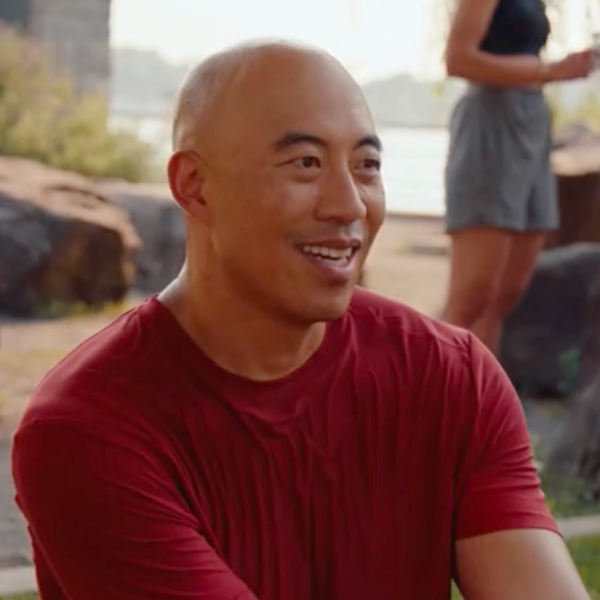 alt="Bald man in a reddish-orange t-shirt smiling outdoors.