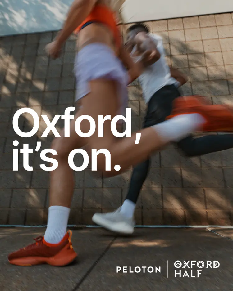 Two runners in action past a brick wall with text reading, Oxford, it's on, Peloton, and Oxford Half.