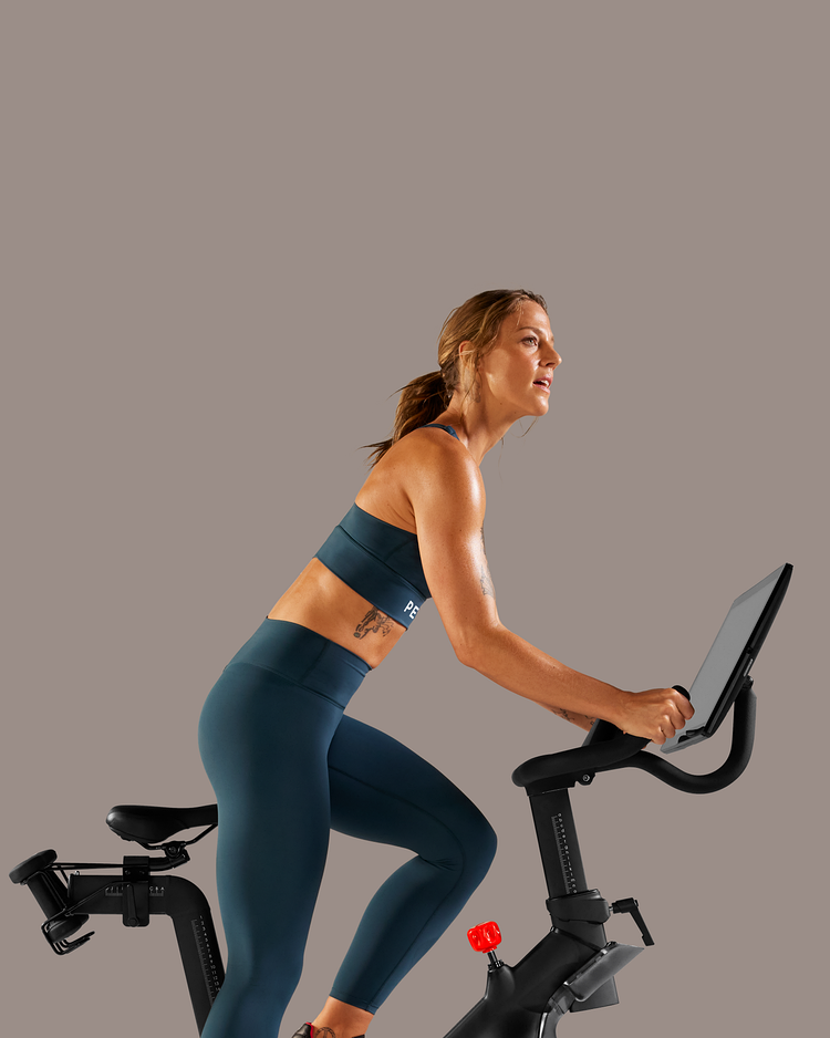 Hände verstellen Touchscreen und Sitzhöhe am Peloton Bike. Füße treten auf dem Peloton Bike in die Pedale. Hand schaltet Peloton Guide ein.
