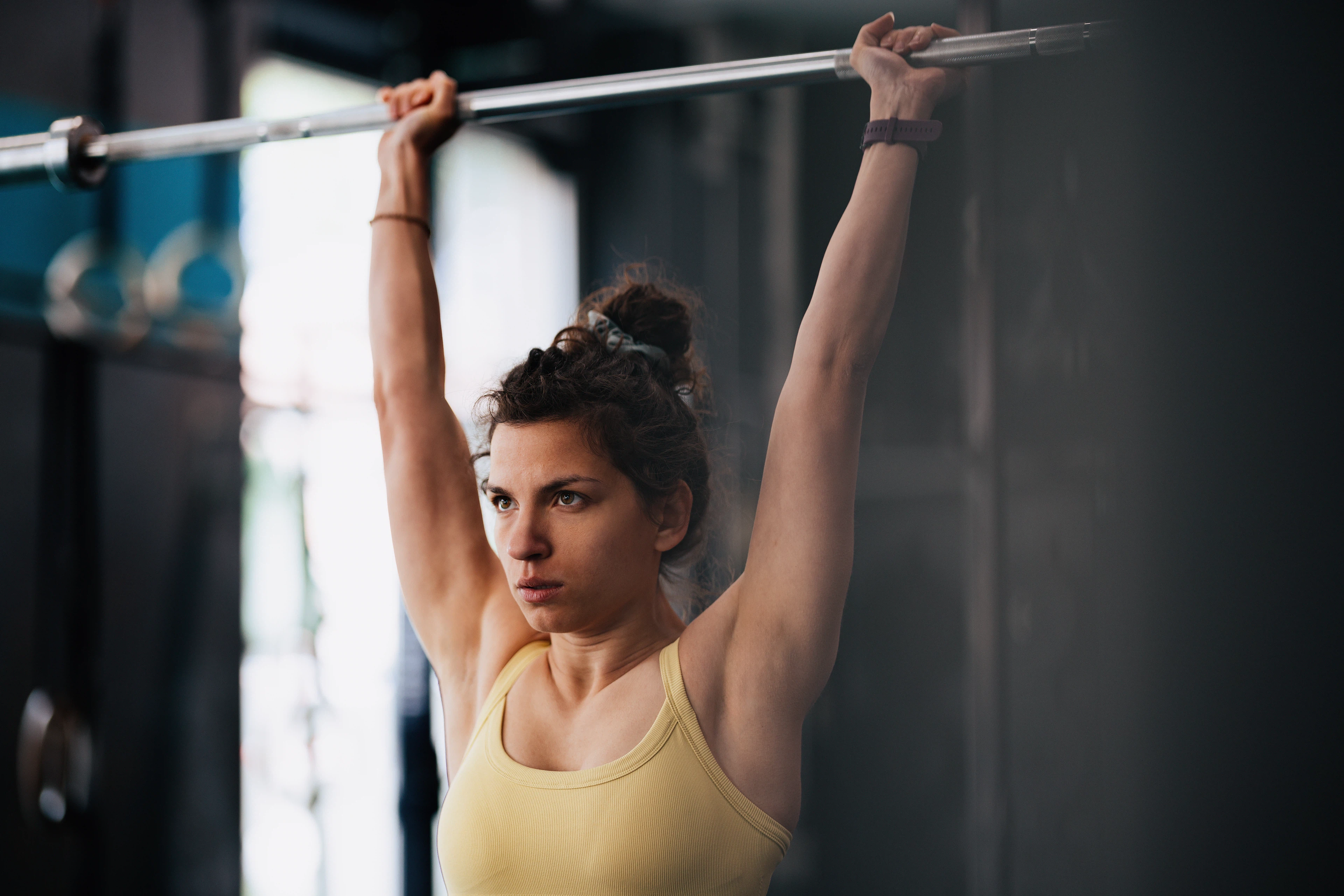 Young woman in a yellow tank top performing an overhead press with a barbell in a gym setting