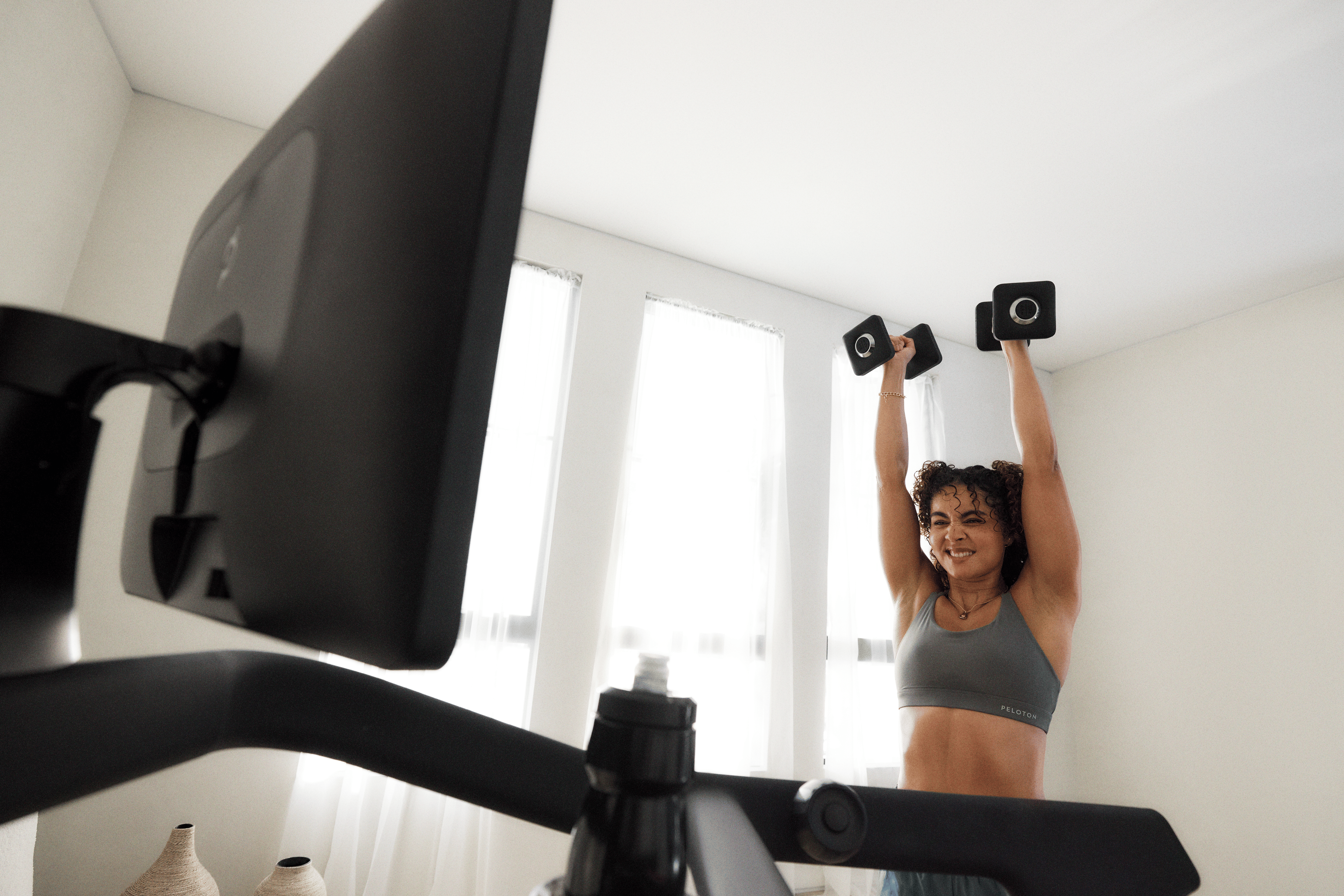 woman doing a shoulder press with dumbbells next to a Peloton Tread