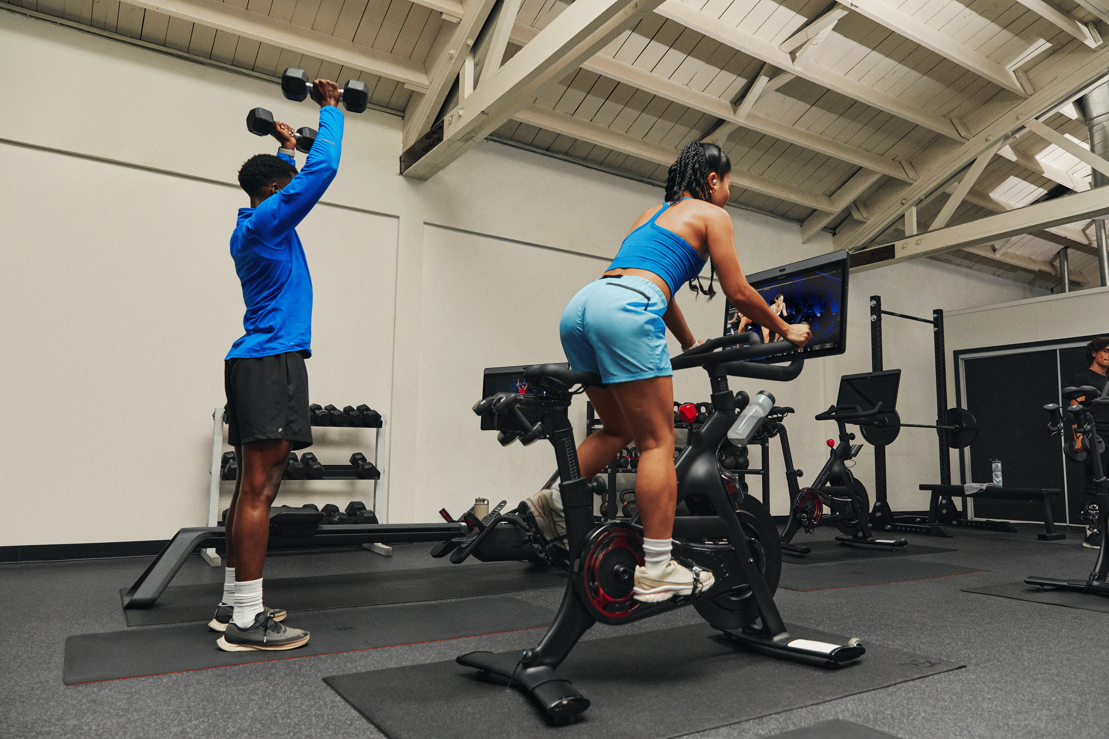 Two friends doing strength and cardio workouts side by side in a gym using a Peloton Bike.