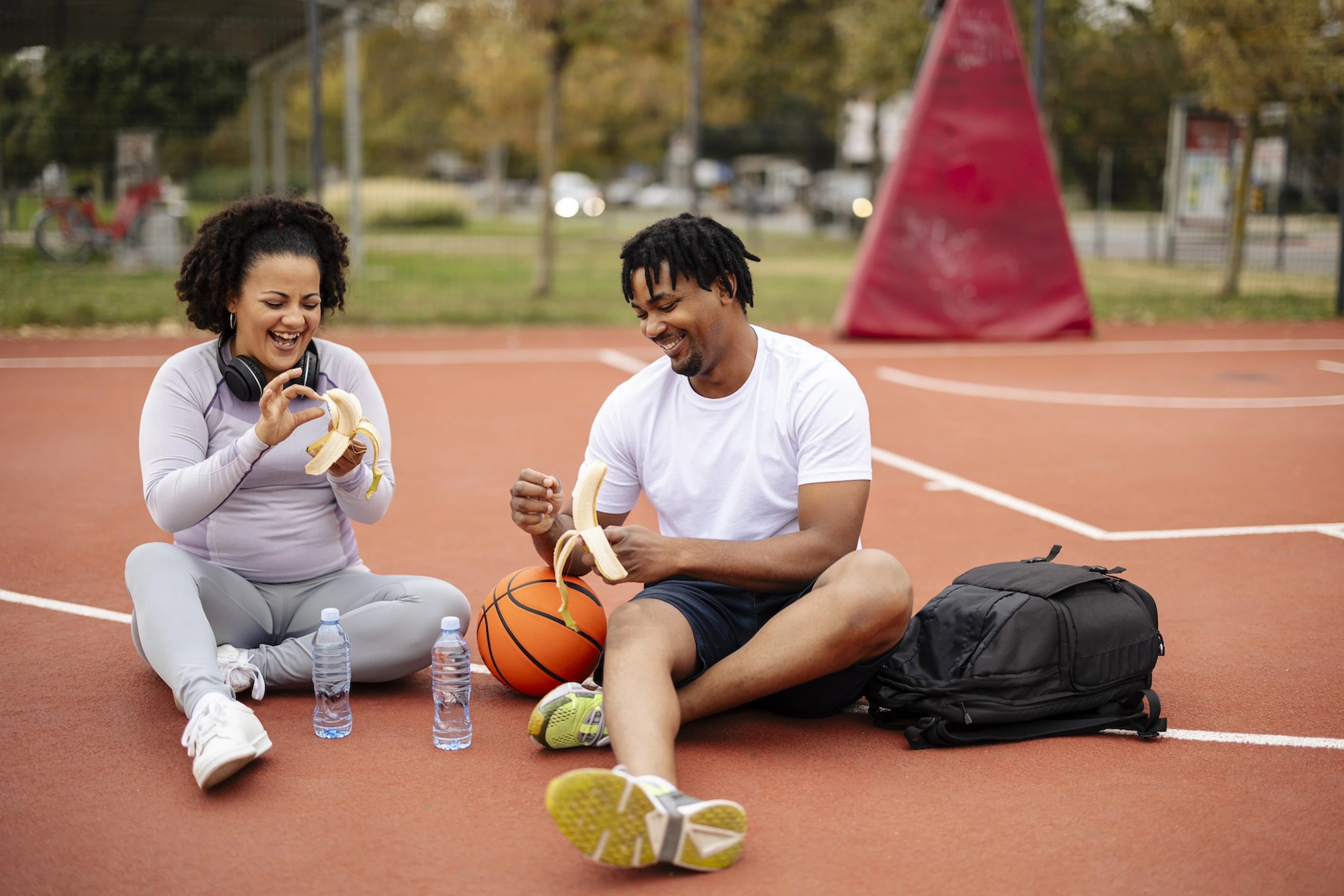 A man and woman sitting on an outdoor basketball court and happily peeling bananas. Learn about the benefits of electrolytes in this article. 