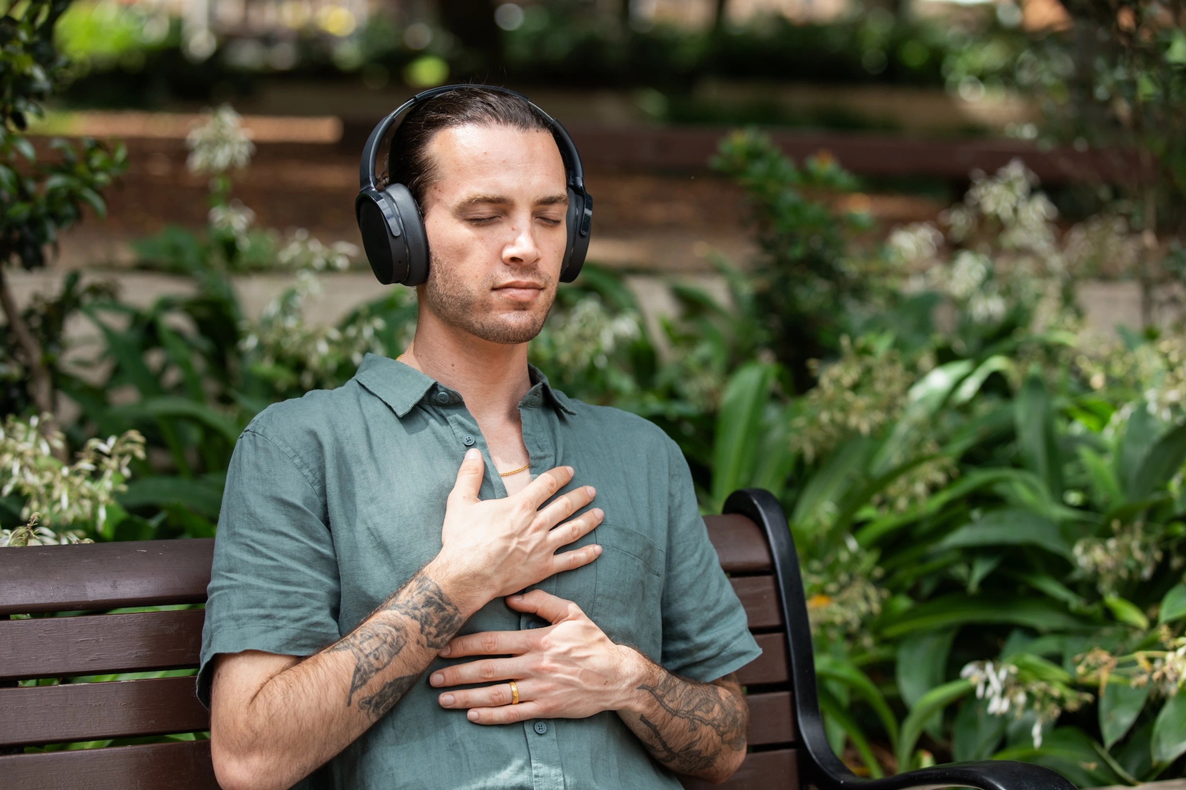 An adult practicing breathwork outside on a park bench. They are listening to headphones and have their hands on their chest.