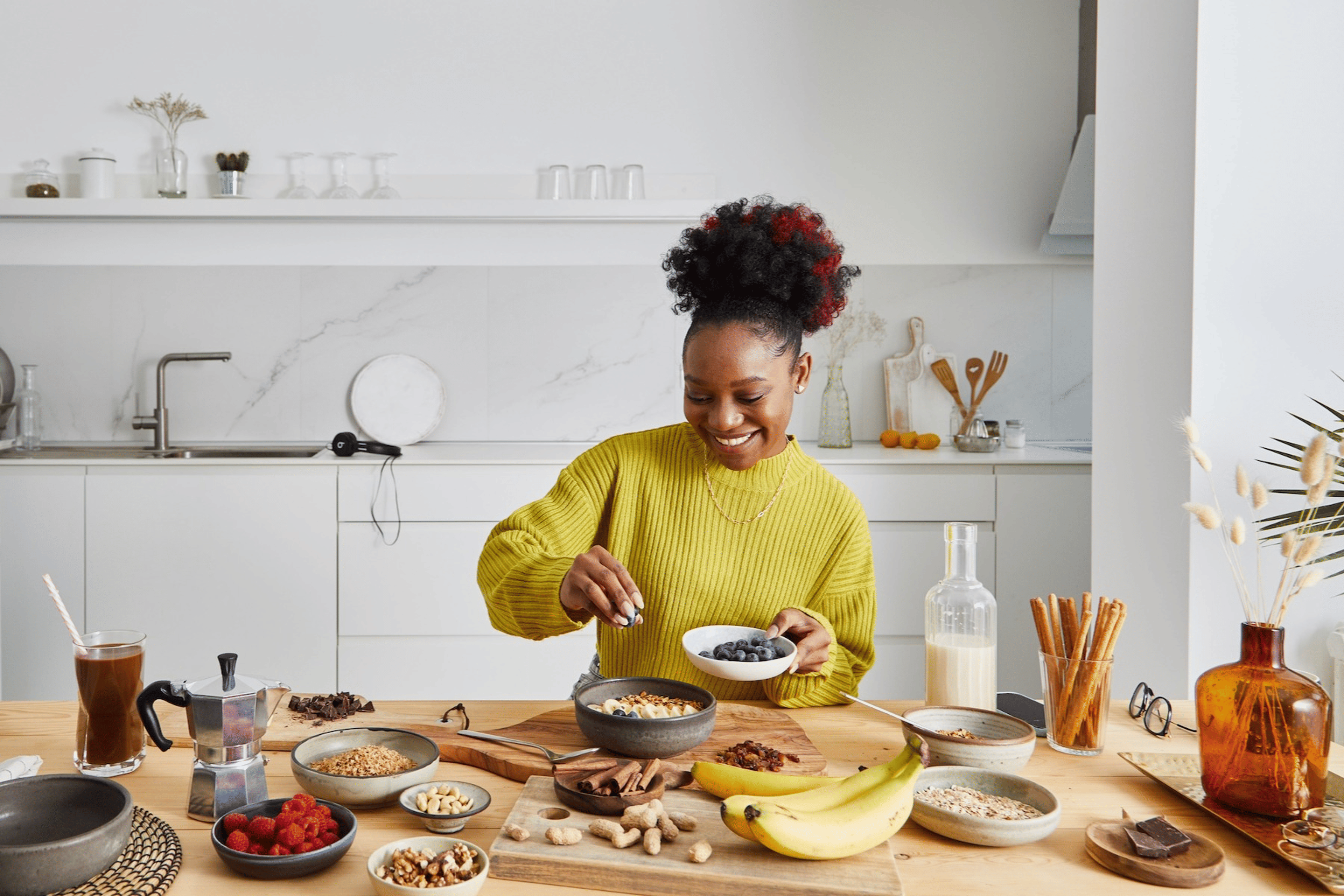 A happy woman at home preparing a healthy breakfast with complex carbohydrates like fruits and nuts.