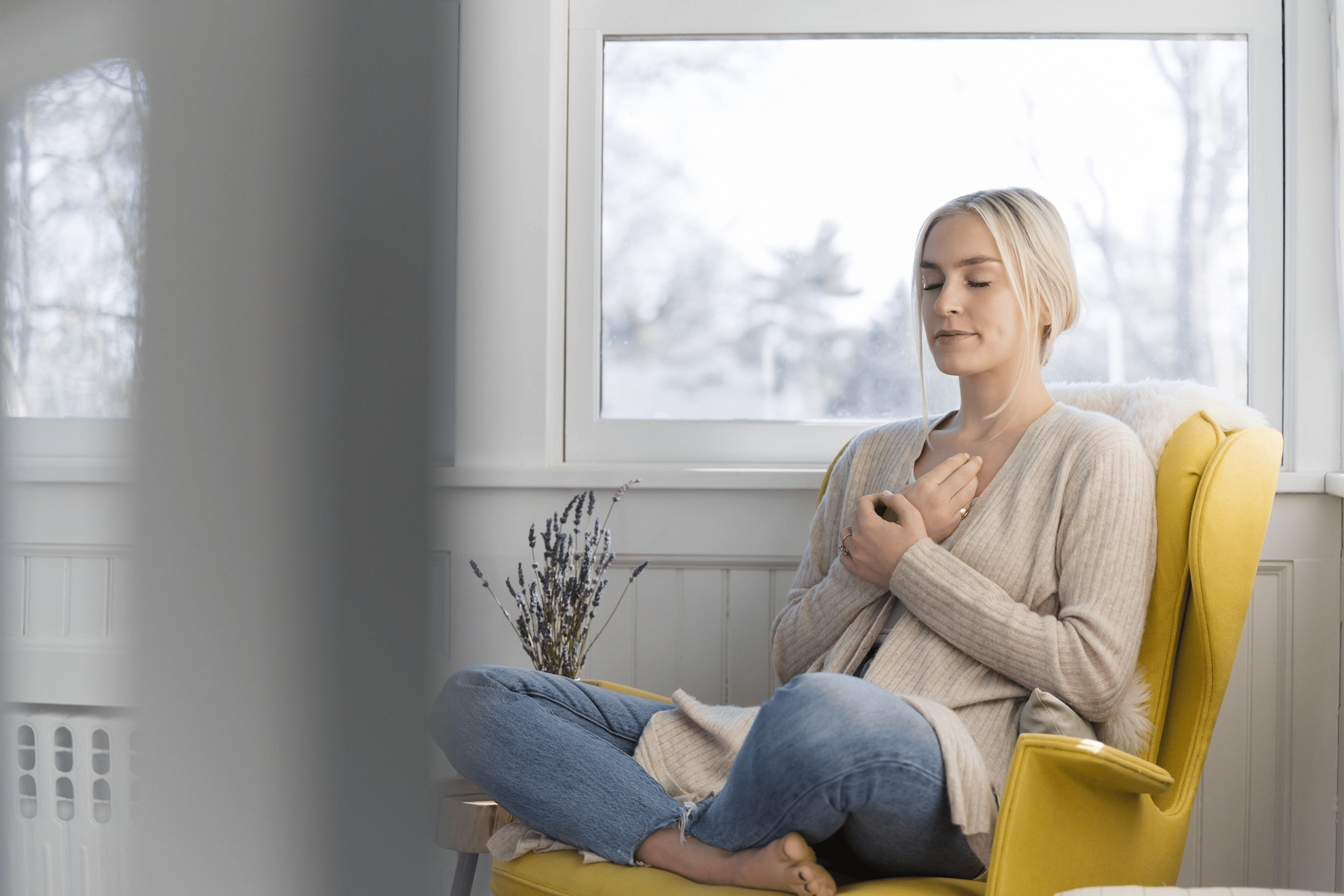 A woman sitting in a chair at home and meditating. Her hands are on her chest, her eyes are closed, and her legs are in a criss-cross position. Learn eight meditation tips in this article.