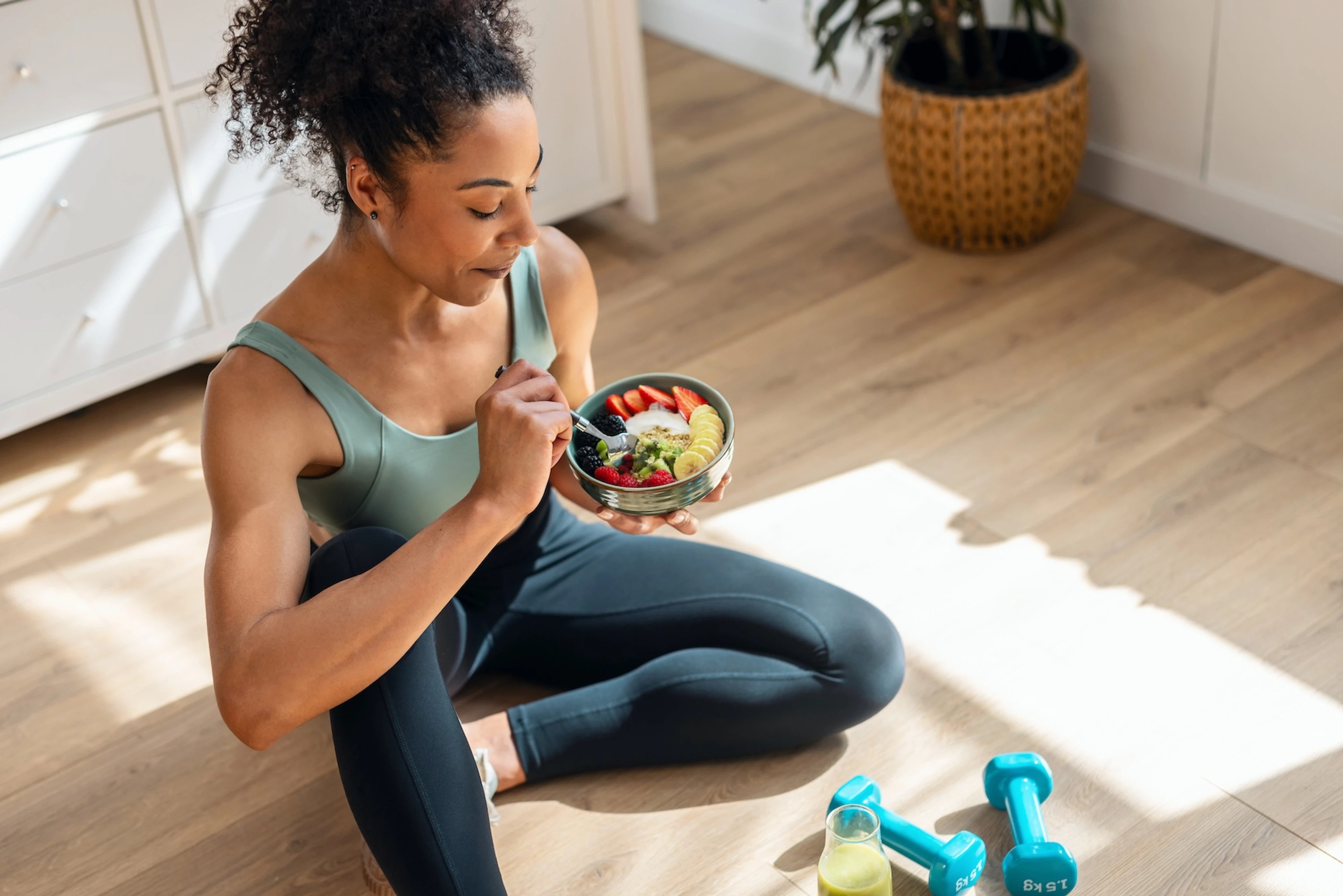 An athlete sitting on the floor at home eating a fruit and yogurt bowl after finishing a dumbbell workout.