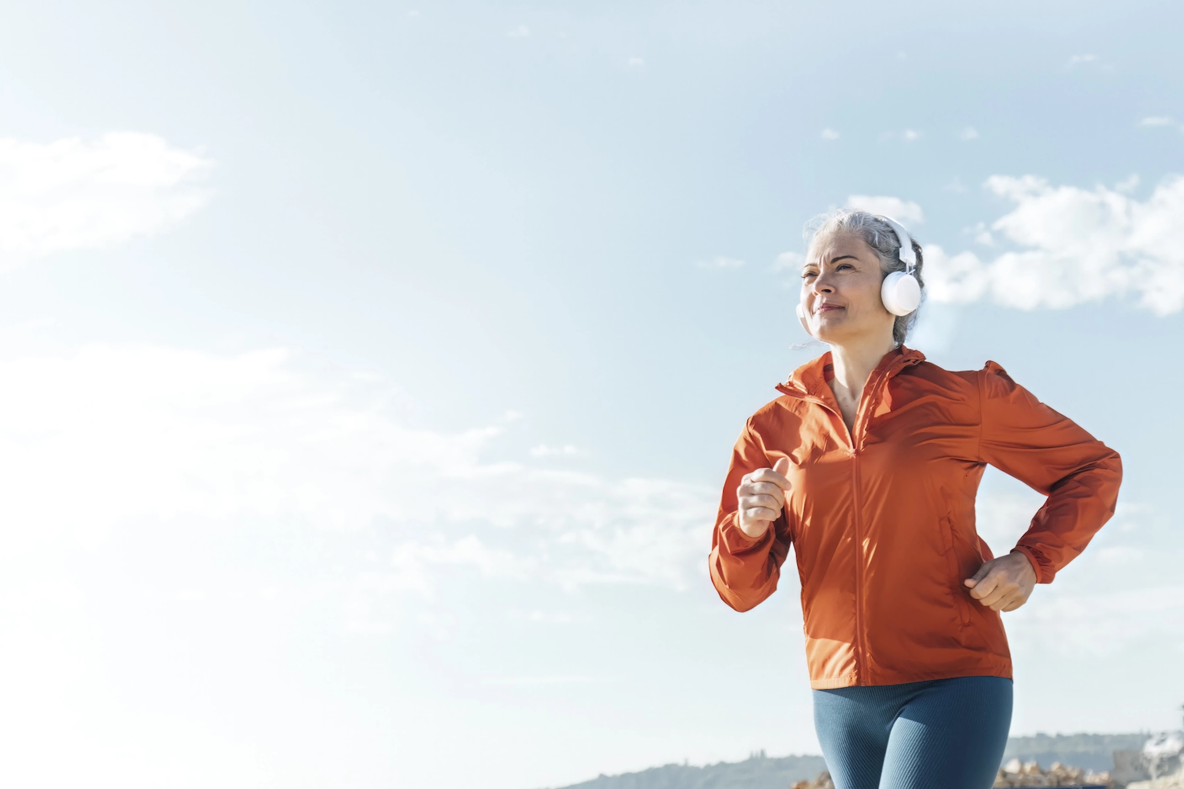 A woman going for an outdoor power walk outside on a sunny day. She's wearing headphones, an orange windbreaker, and blue leggings.