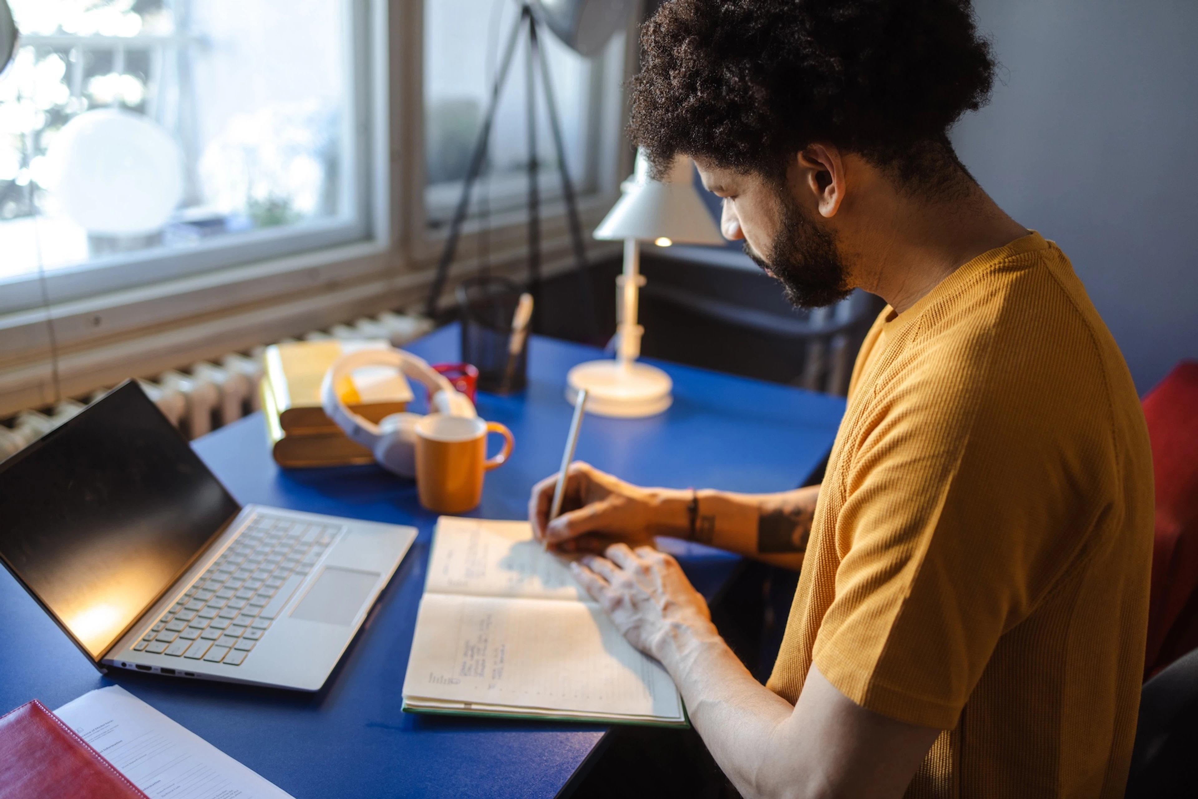 A man being productive at home. He's writing something down in his notebook while sitting at his desk.