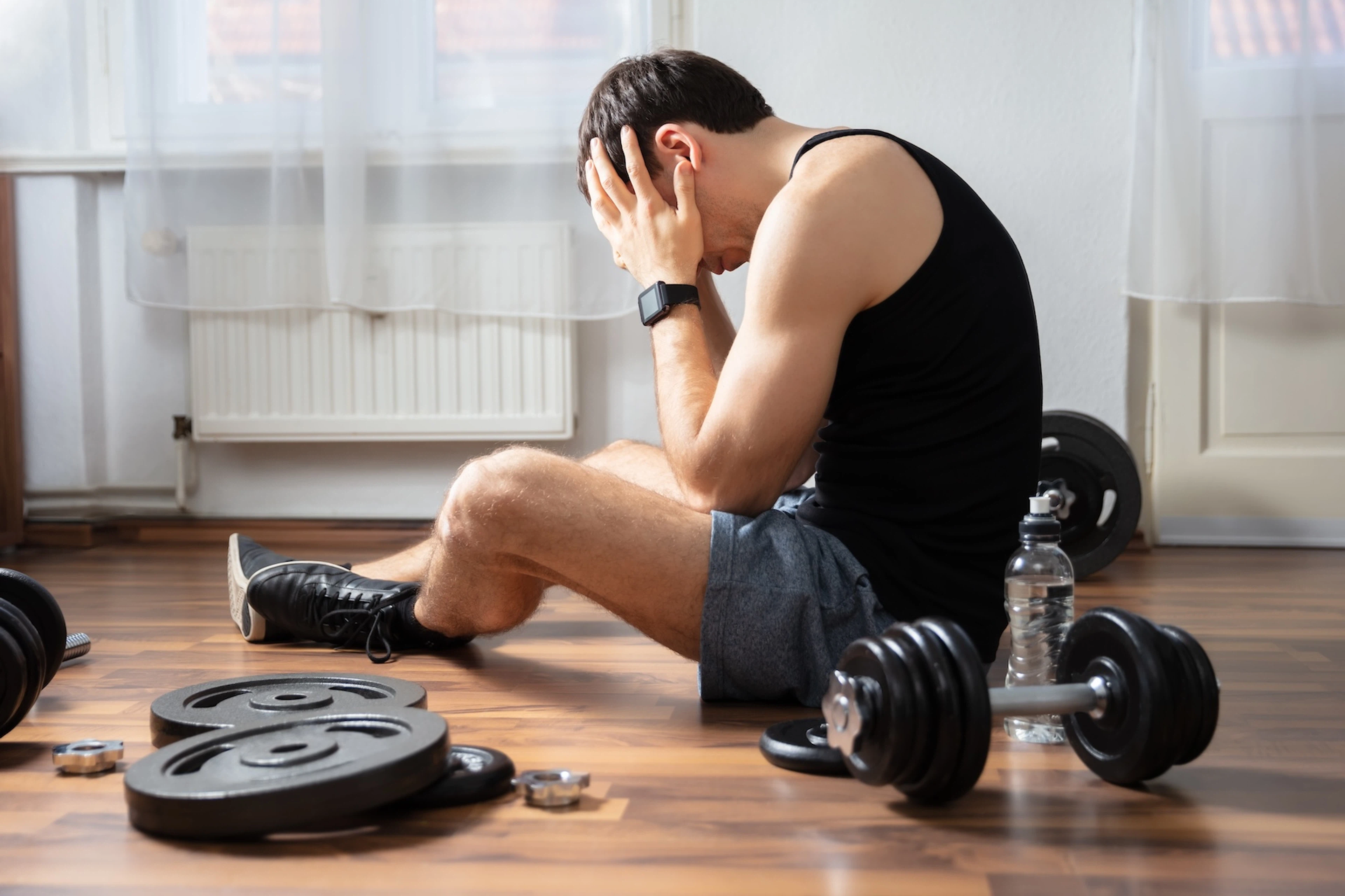 A man dealing with exercise burnout. His head is in his hands and he's feeling mentally drained, surrounded by dumbbells and weights.