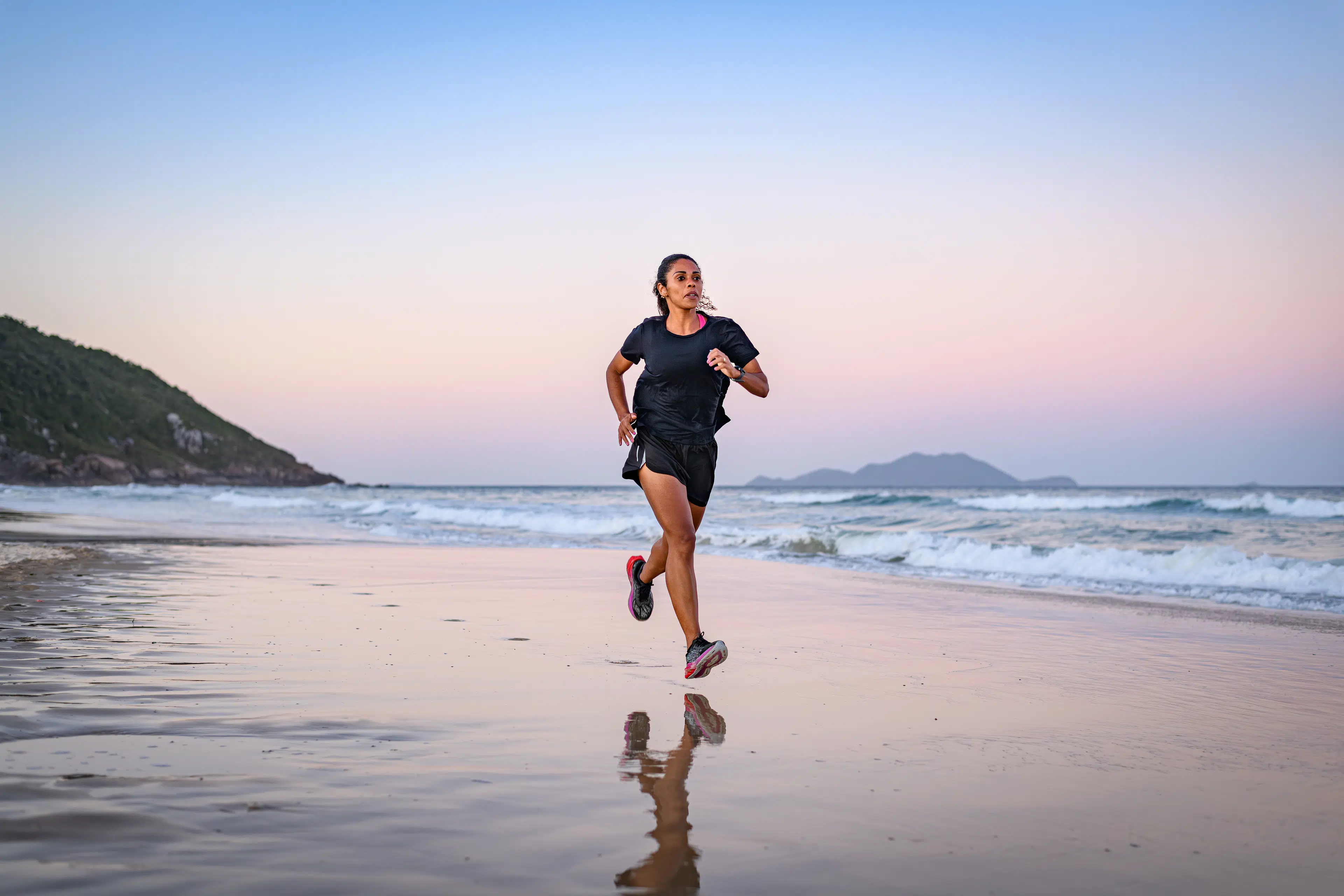 Woman running on the beach at sunset in sneakers and a black outfit.