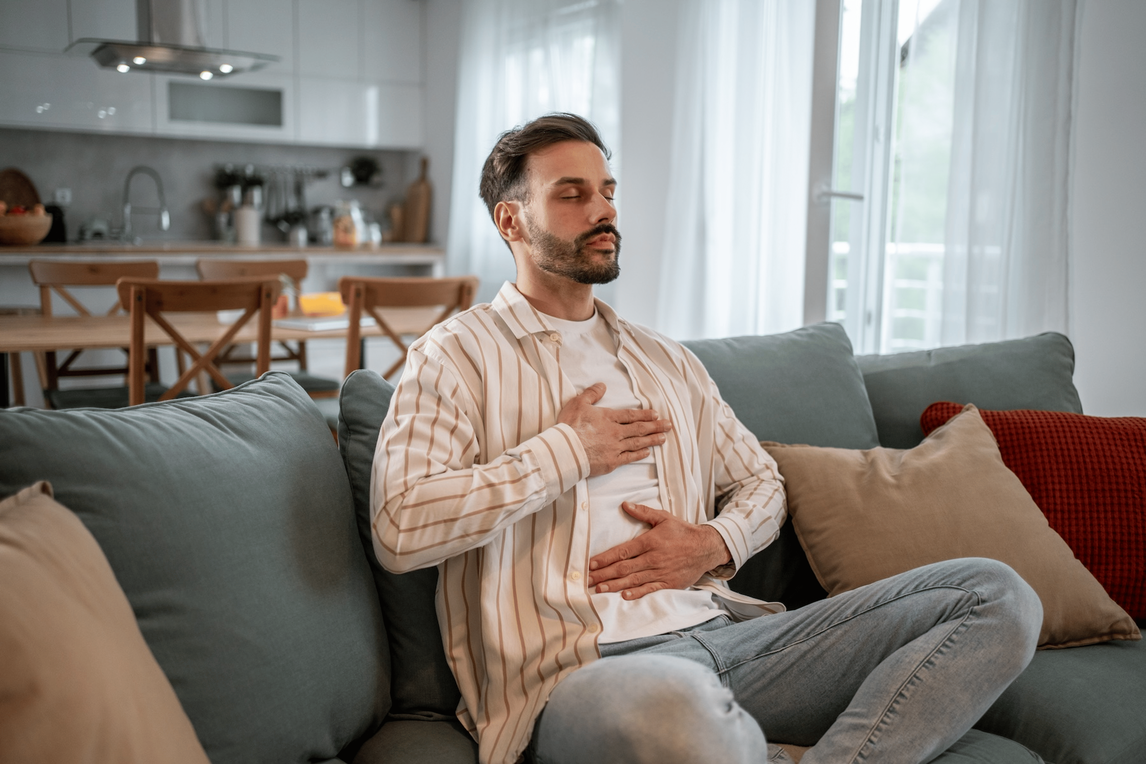A man sitting on a cough at home practicing belly breathing. His eyes are closed and he has one hand on his chest and the other on his belly. 
