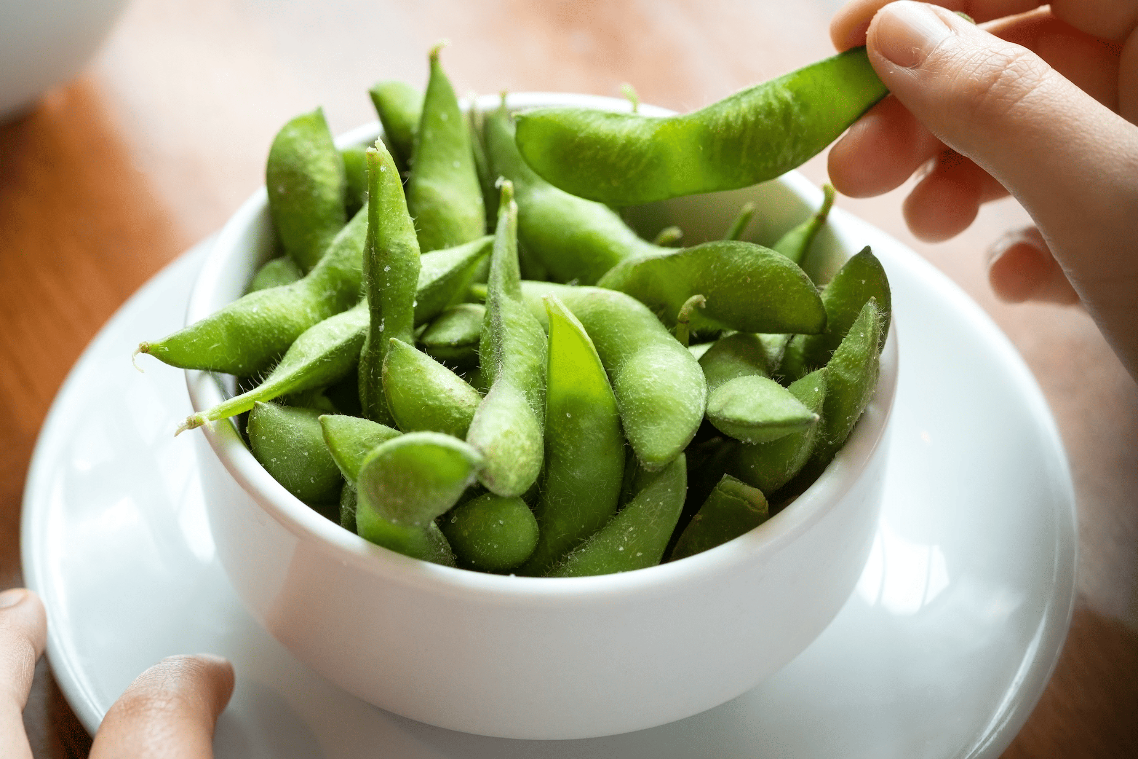 Close-up photo of a hand eating from a bowl of salted edamame.