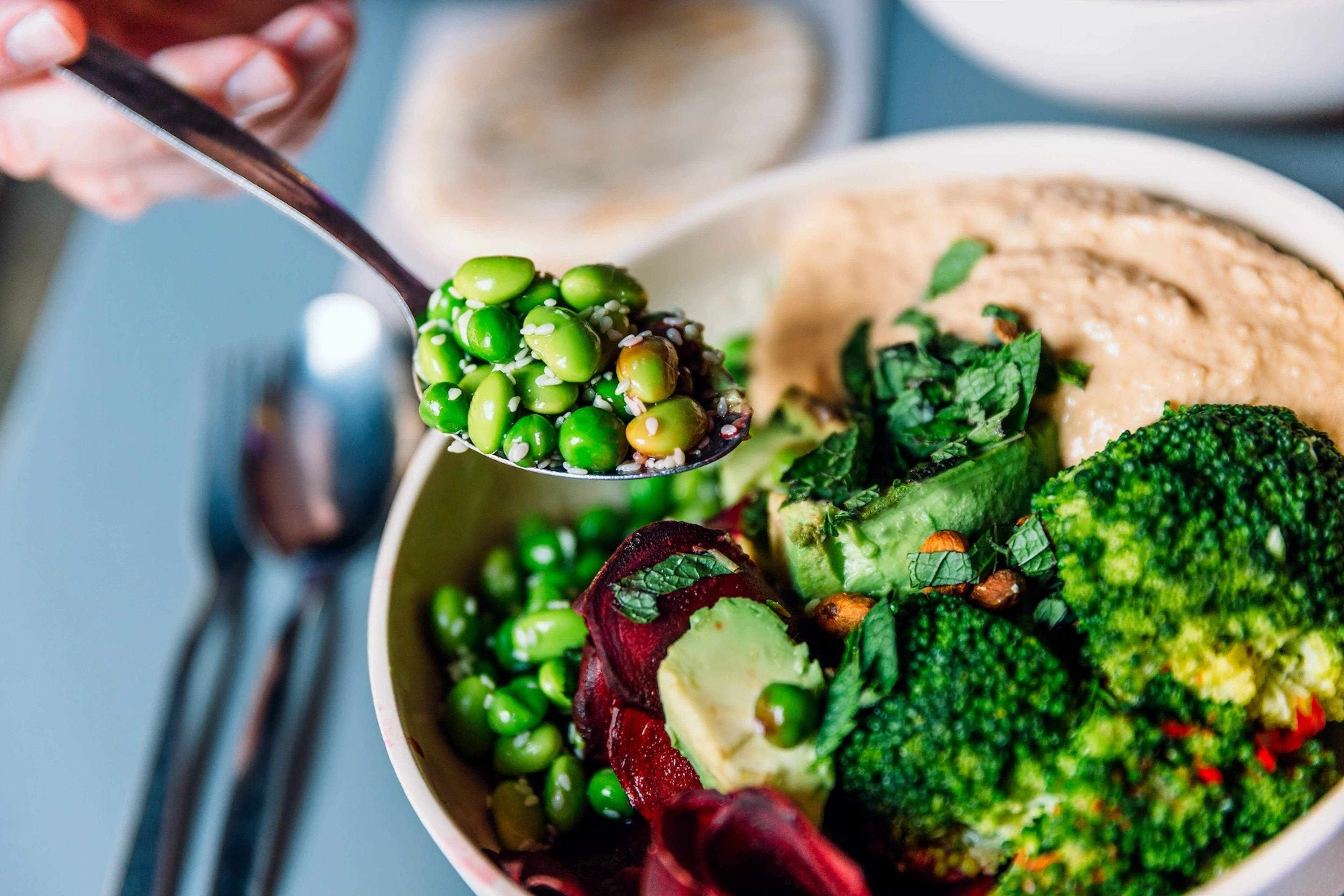 A spoon of edamame (a non-meat protein source) being eaten from a bowl with broccoli, beetroot, avocado, nuts, and hummus.