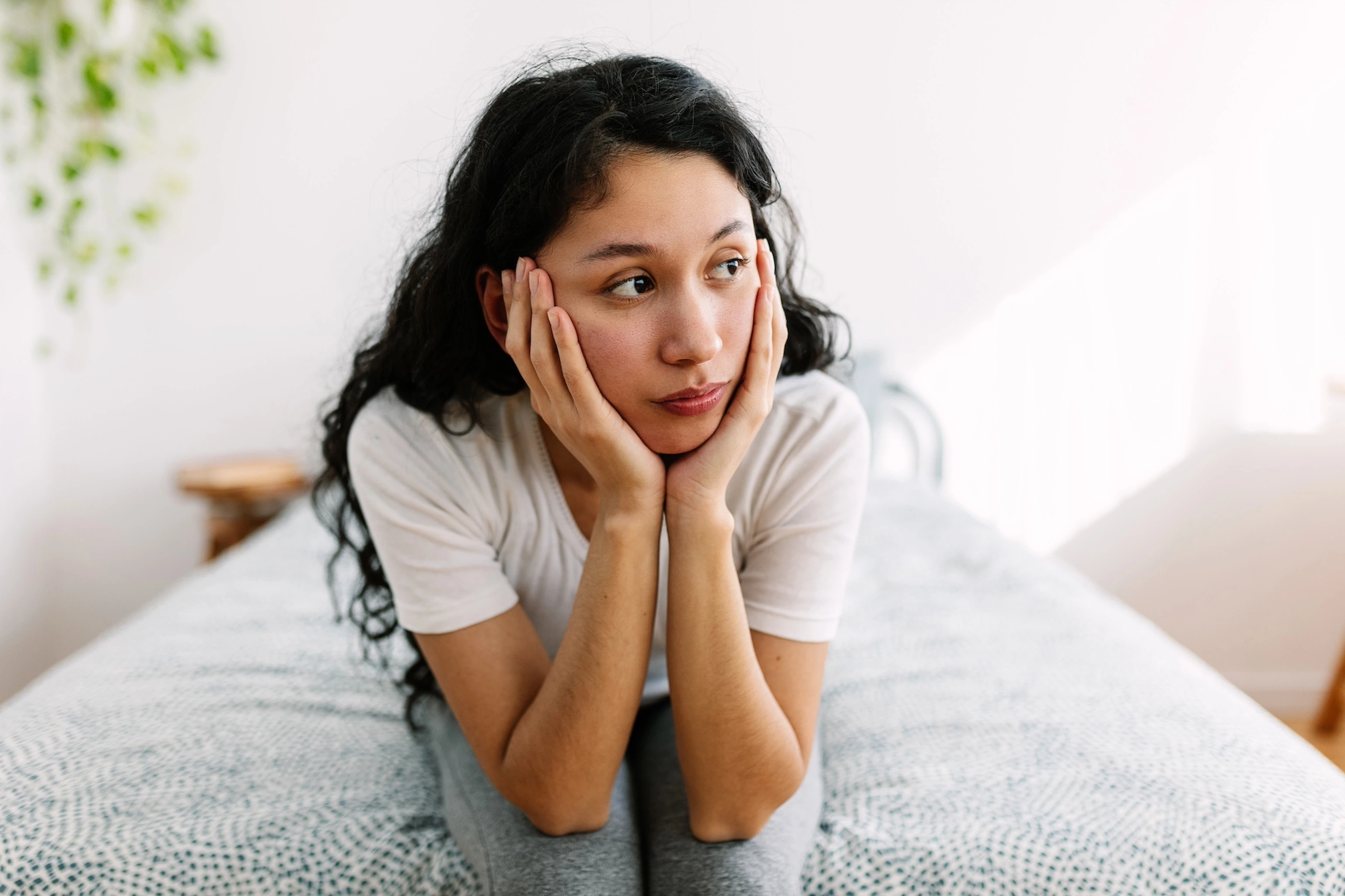 A young woman experiencing the Sunday scaries. She's sitting on her bed with her head in her hands and looking away from the camera.