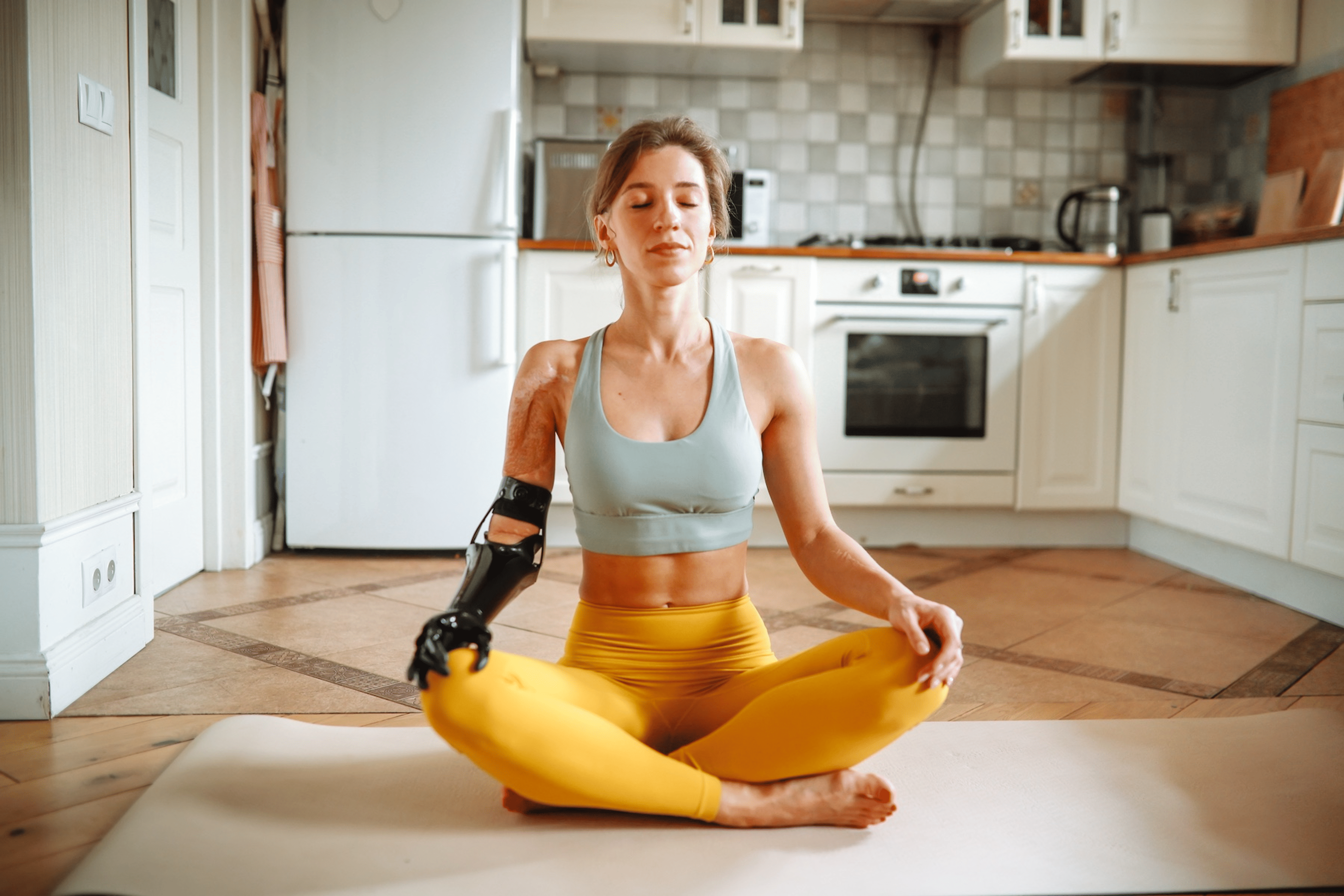 A woman meditating on her yoga mat at home in her kitchen. She's sitting in lotus position, closing her eyes, and has her hands resting on her knees.
