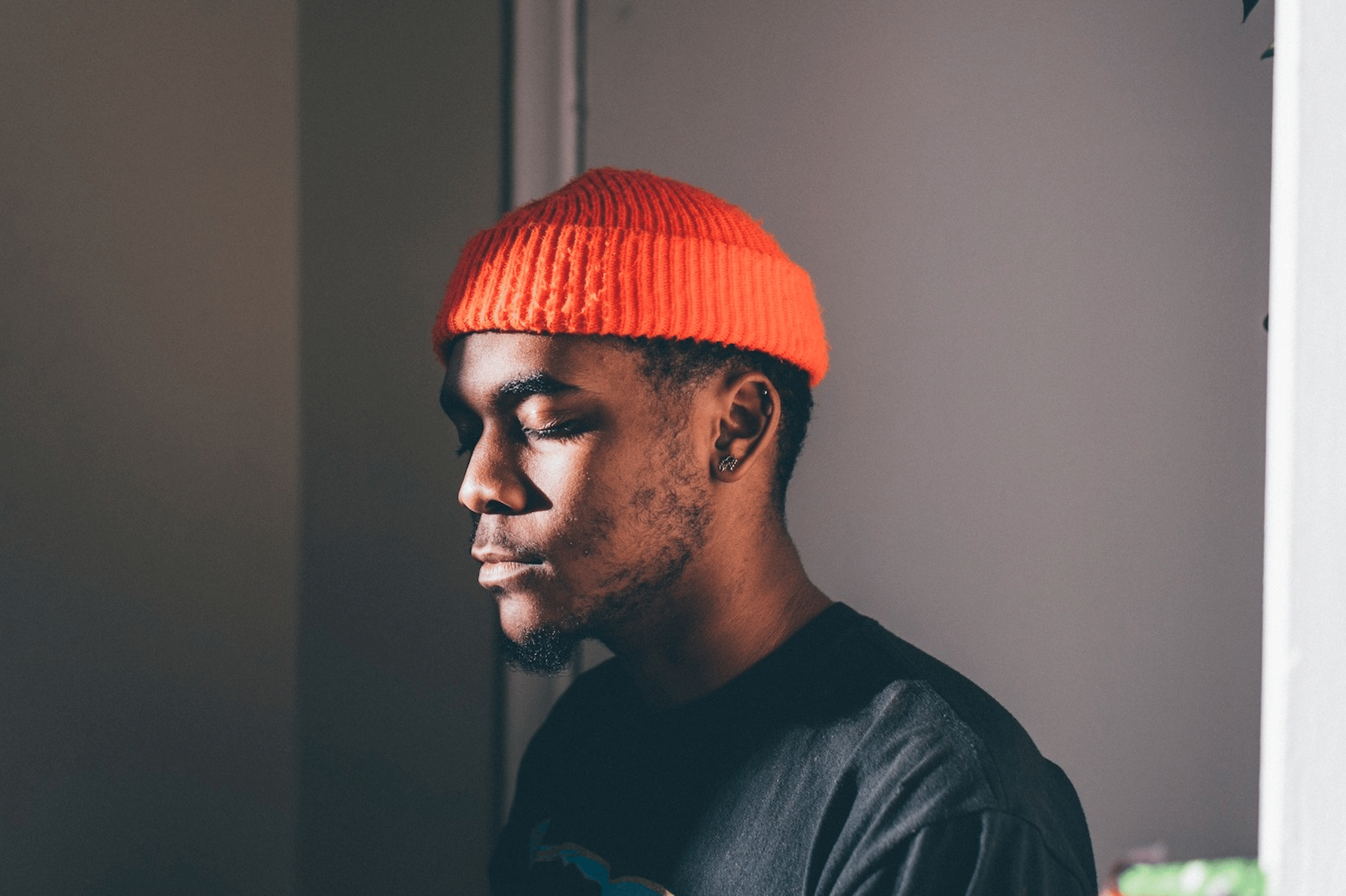 A man wearing an orange beanie sitting in the light while closing his eyes and practicing visualization meditation.