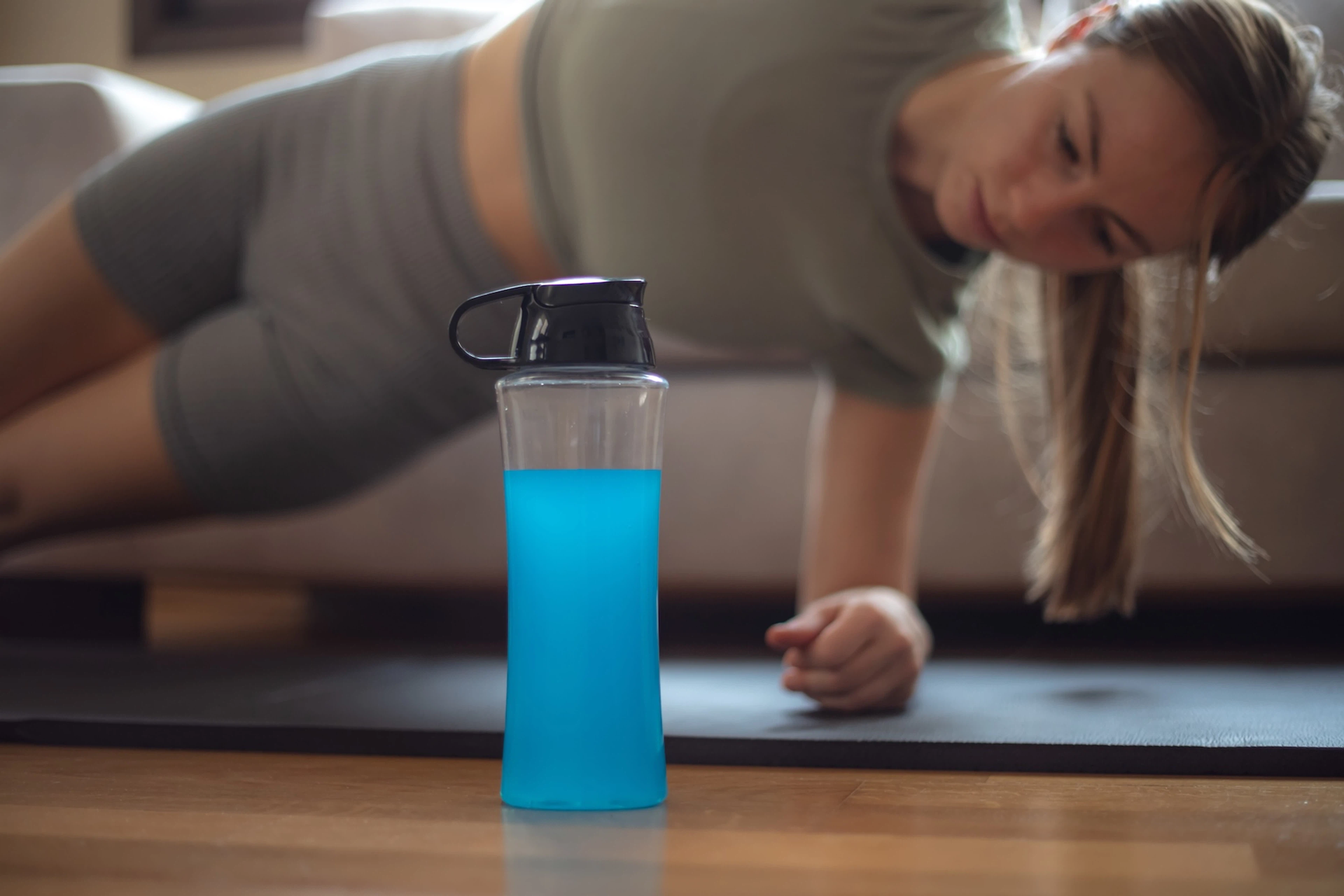 A blue sports drink in the foreground with a woman doing a side plank on a workout mat in the background.