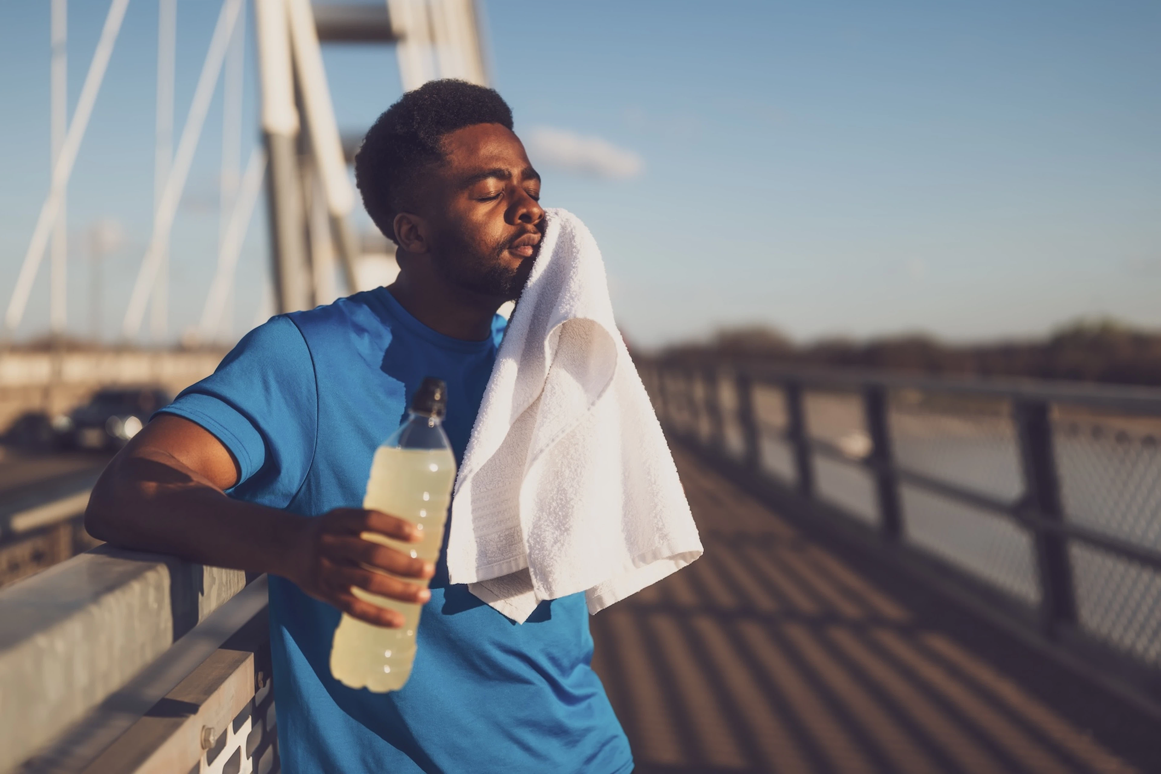 A man experiencing tooth pain after running. He is holding a white towel up to his jaw and closing his eyes while resting and holding a sports drink.
