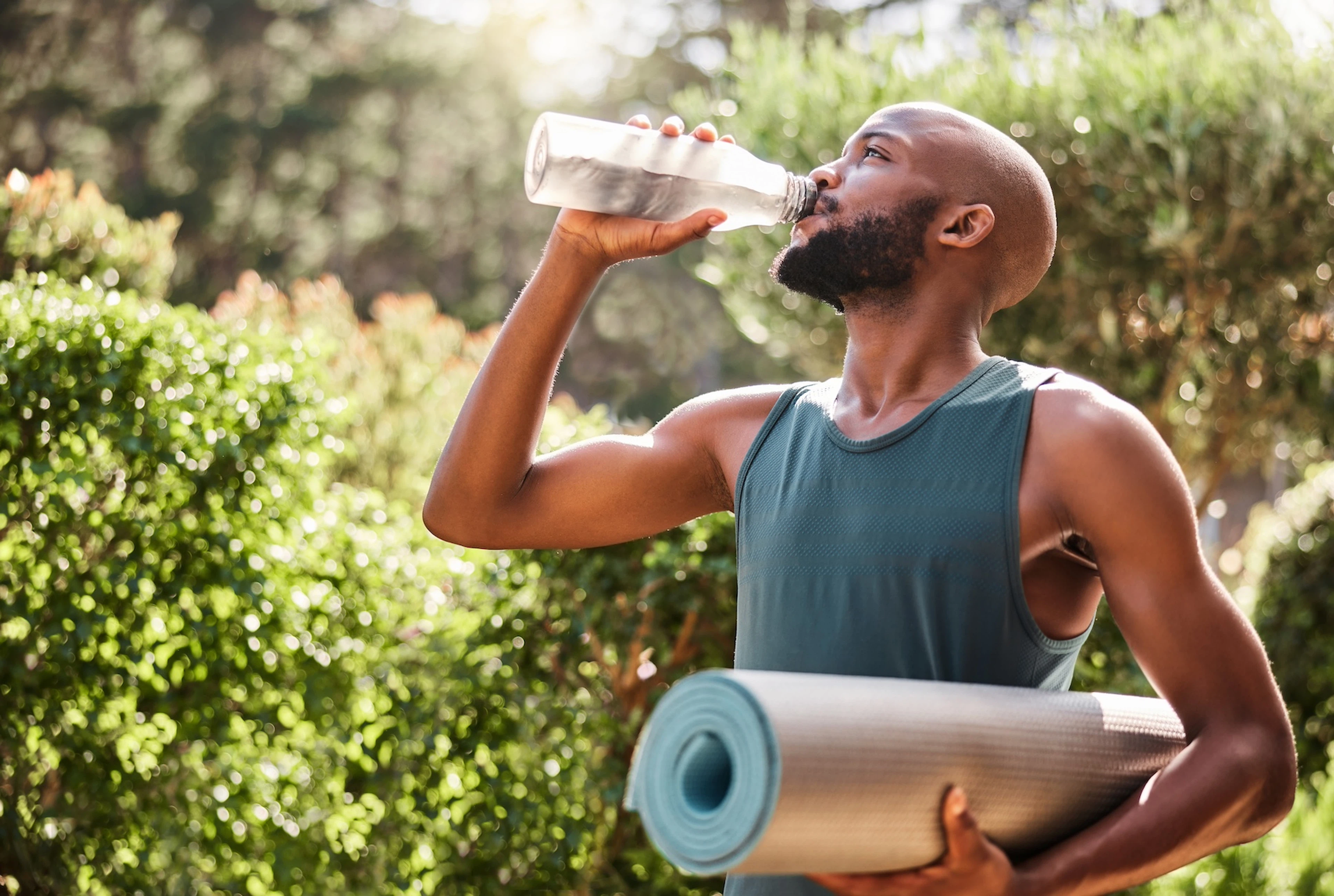 A man carrying a yoga mat under his arm and drinking from a water bottle with the other. Learn how to rehydrate in this article.