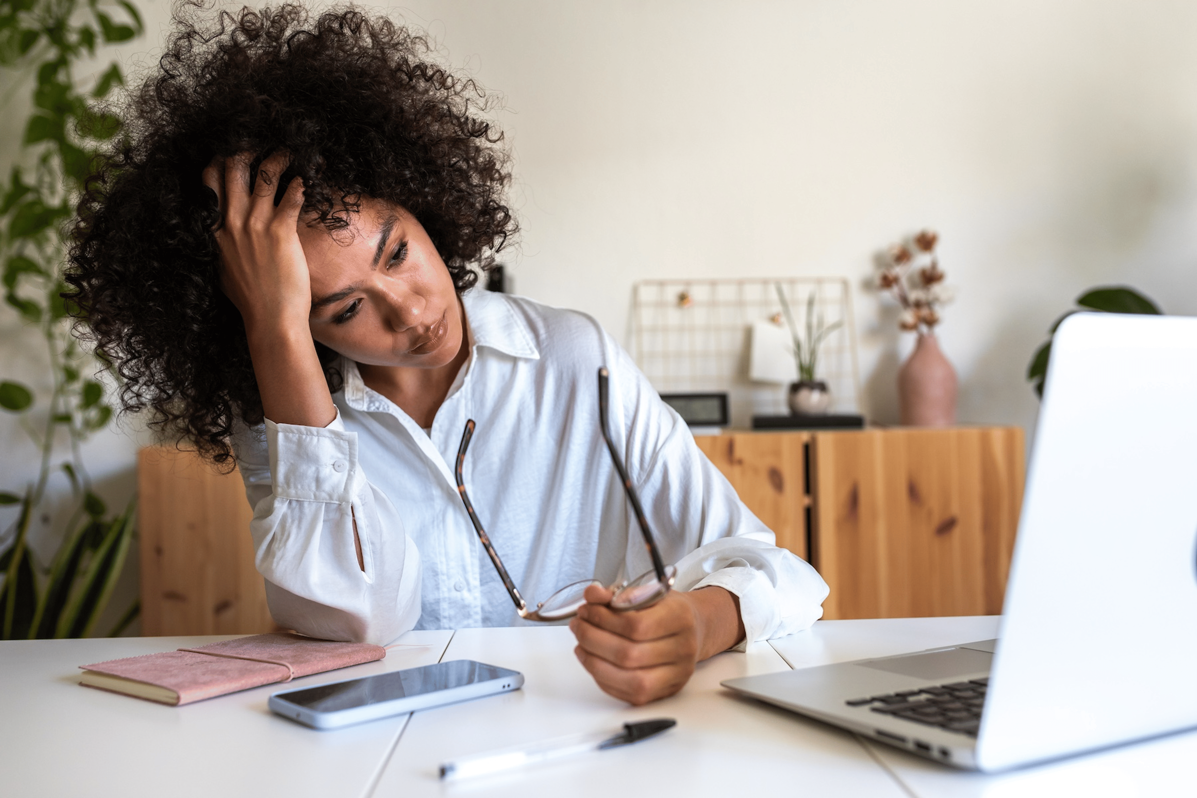 A mentally exhausted woman sitting at her desk in front of her laptop. She's holding her head in one hand and her glasses in another. She looks stressed out and tired.