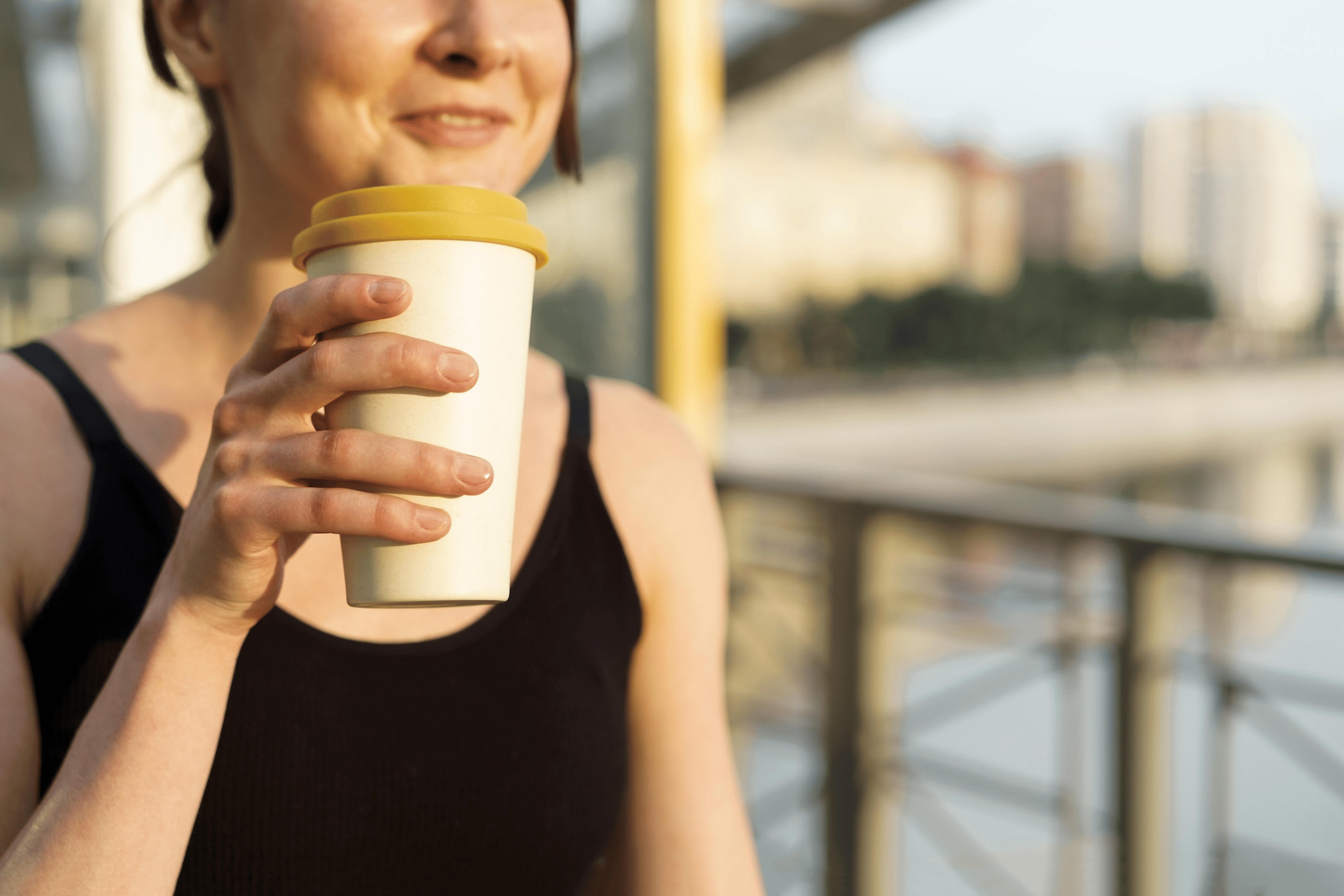 A close-up photo of a woman drinking coffee after a workout. She's standing outside and holding a reusable to-go cup.
