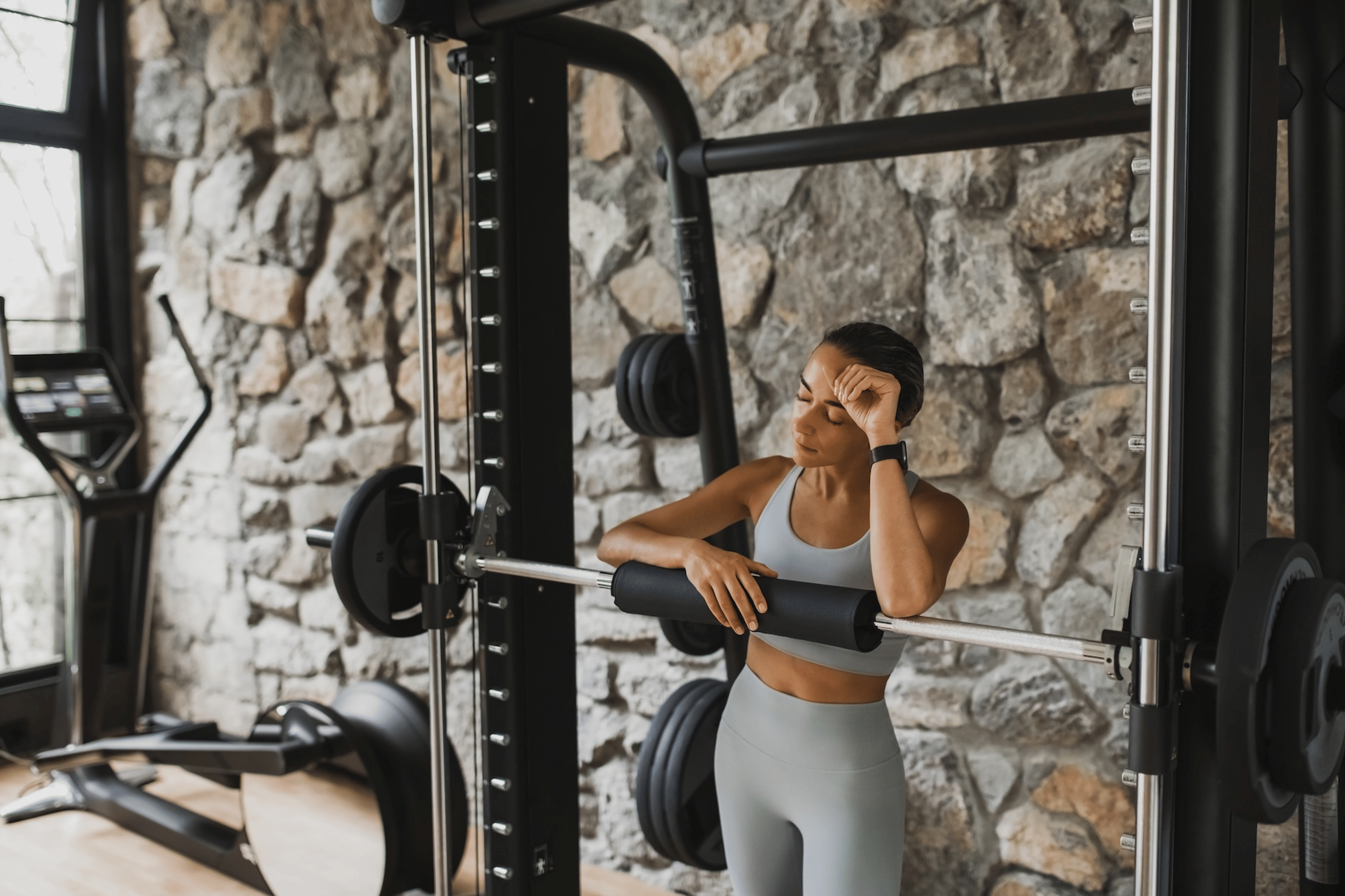 A woman in the gym near a barbell set. Her eyes are closed and her hand is on her head as she tries to work out with a headache.