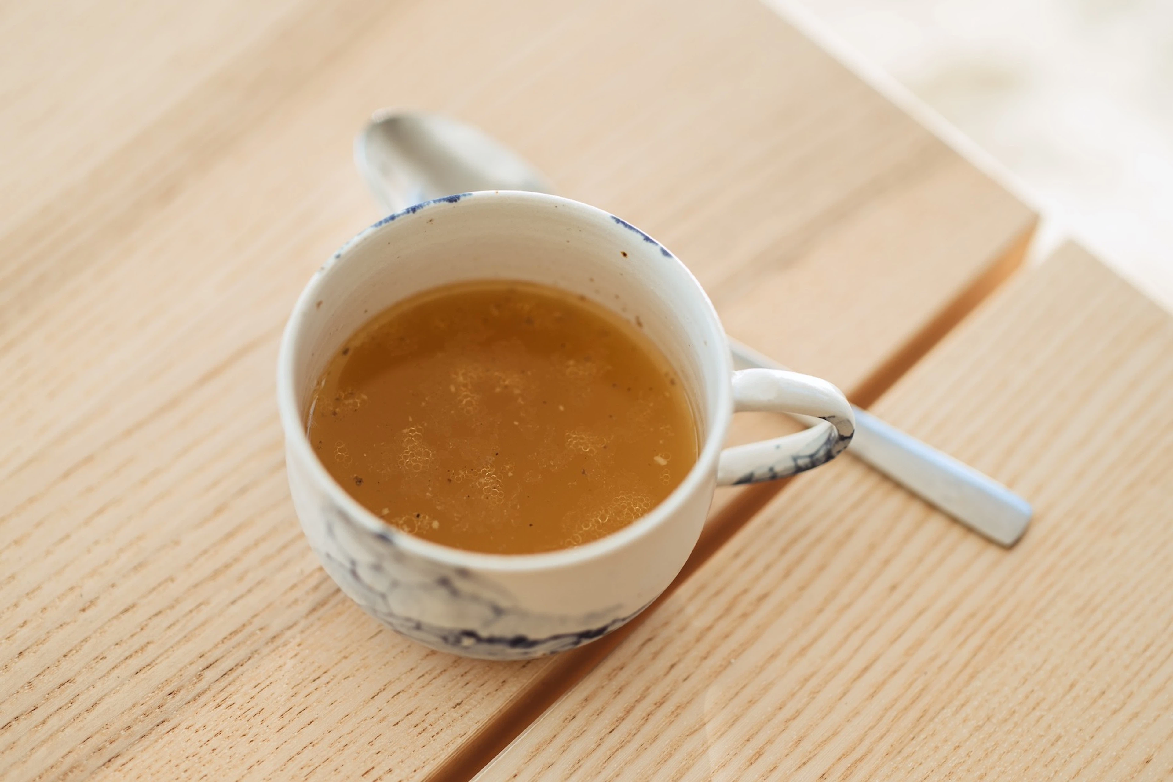 A mug of bone broth, a hydrating food, next to a spoon resting on a table.