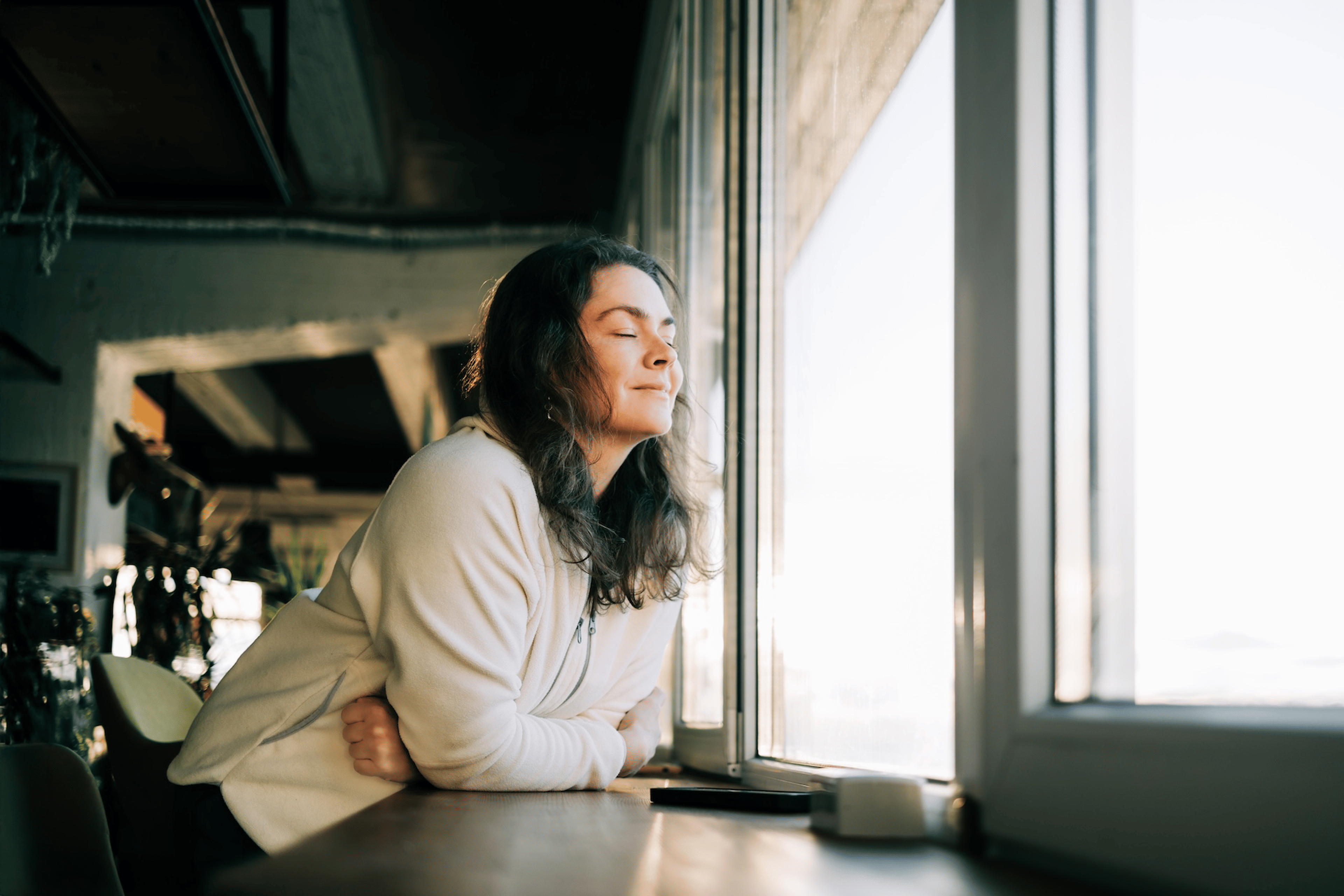 A woman leaning on a counter near a window, closing her eyes and taking time to be mindful.