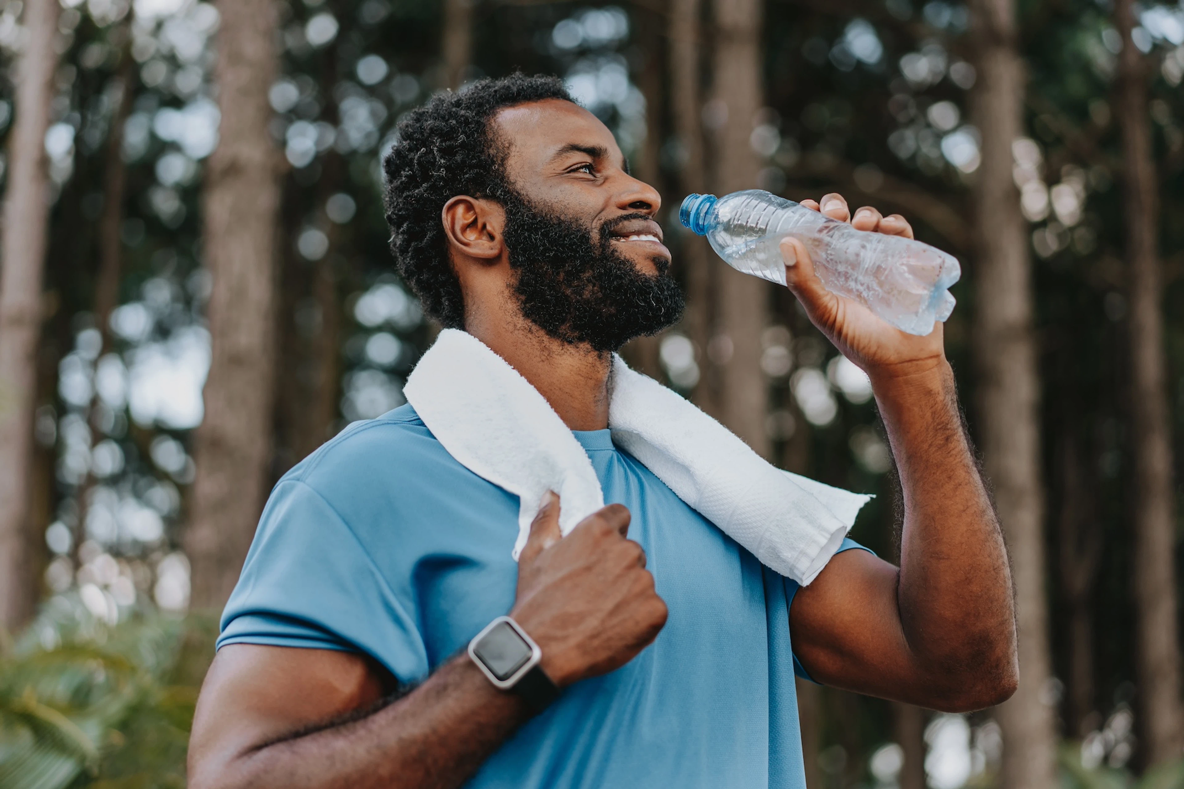 A man drinking from a water bottle after a workout outdoors. He is wearing a blue shirt and has a white towel around his neck.
