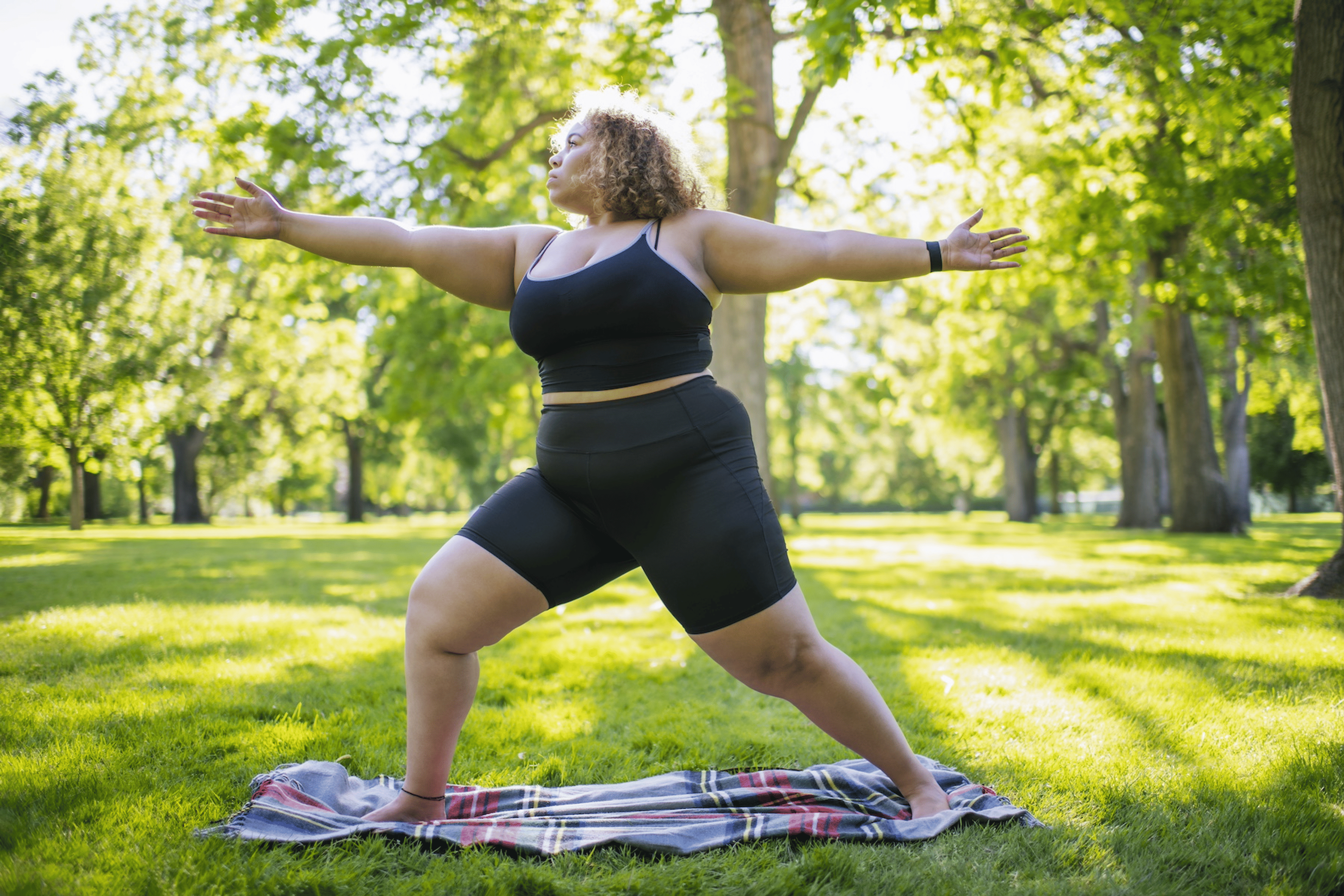 A woman working out barefoot. She is in Warrior 2 pose while practicing yoga on a blanket in a park.
