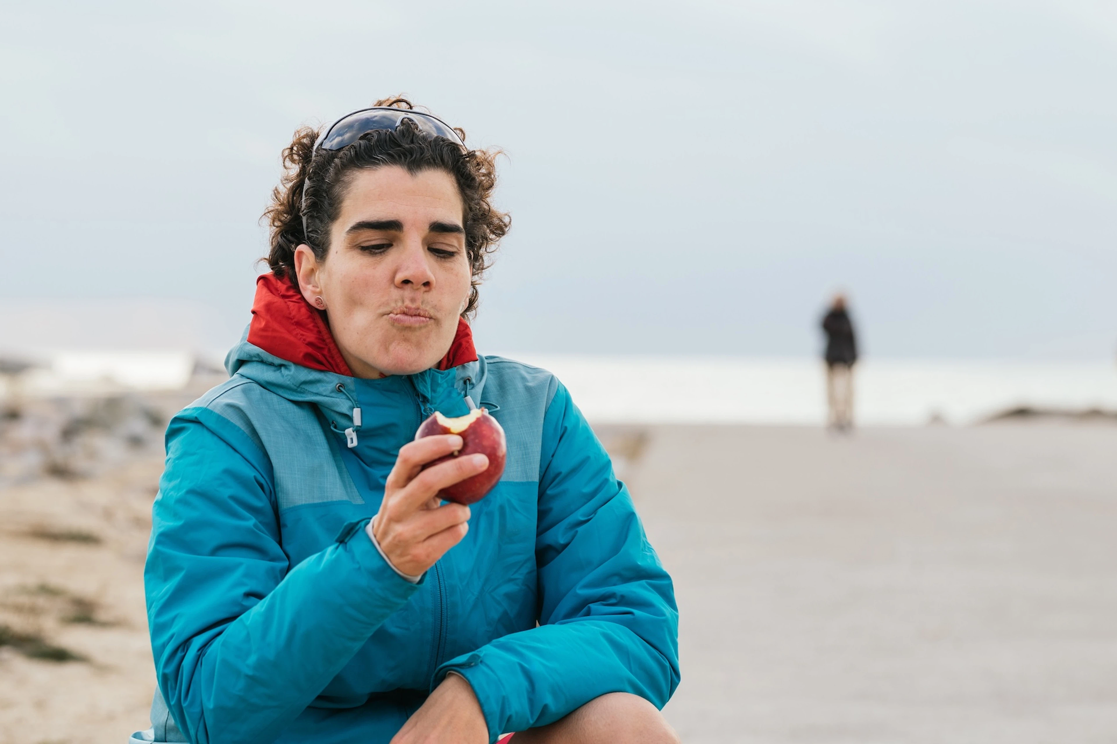 A runner eating an apple shortly before, during, or after a run on the beach. She is preventing runger by taking in some fuel.