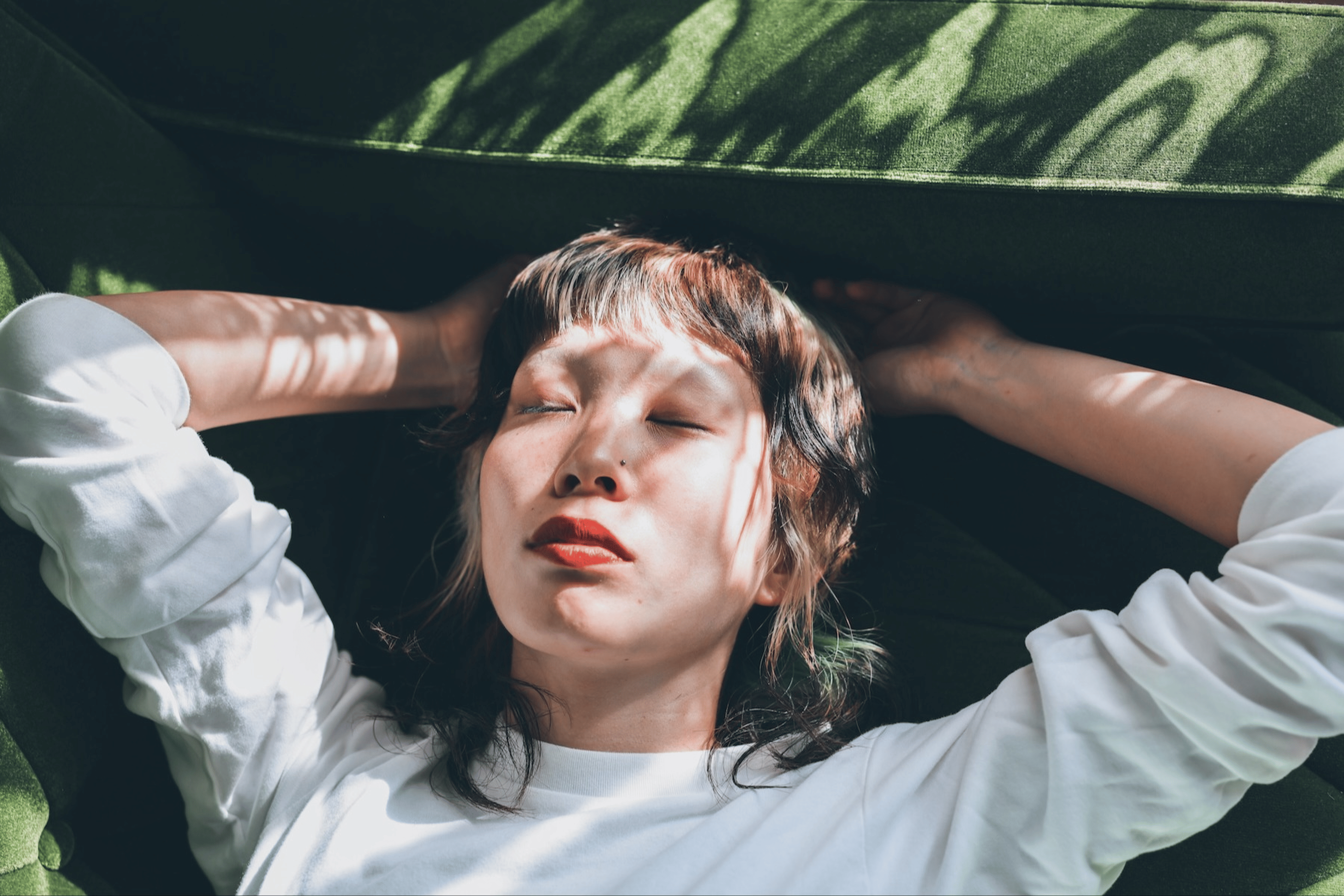 A woman relaxing at home and practicing progressive muscle relaxation. She's lying on a sunny sofa with her eyes closed and hands behind her head.