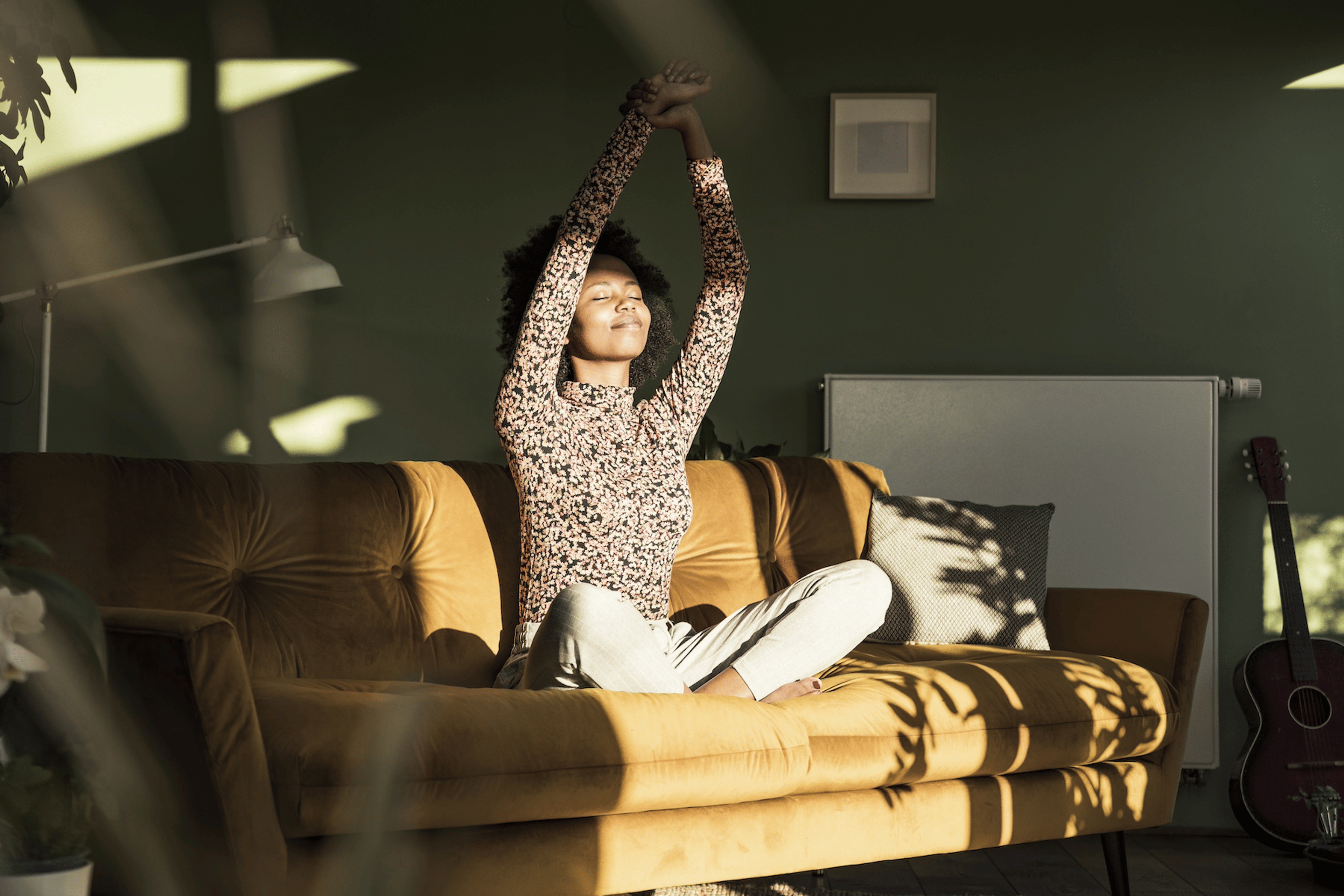 A woman stretching her arms overhead while closing her eyes and smiling. She is sitting on the couch in the sunlight.