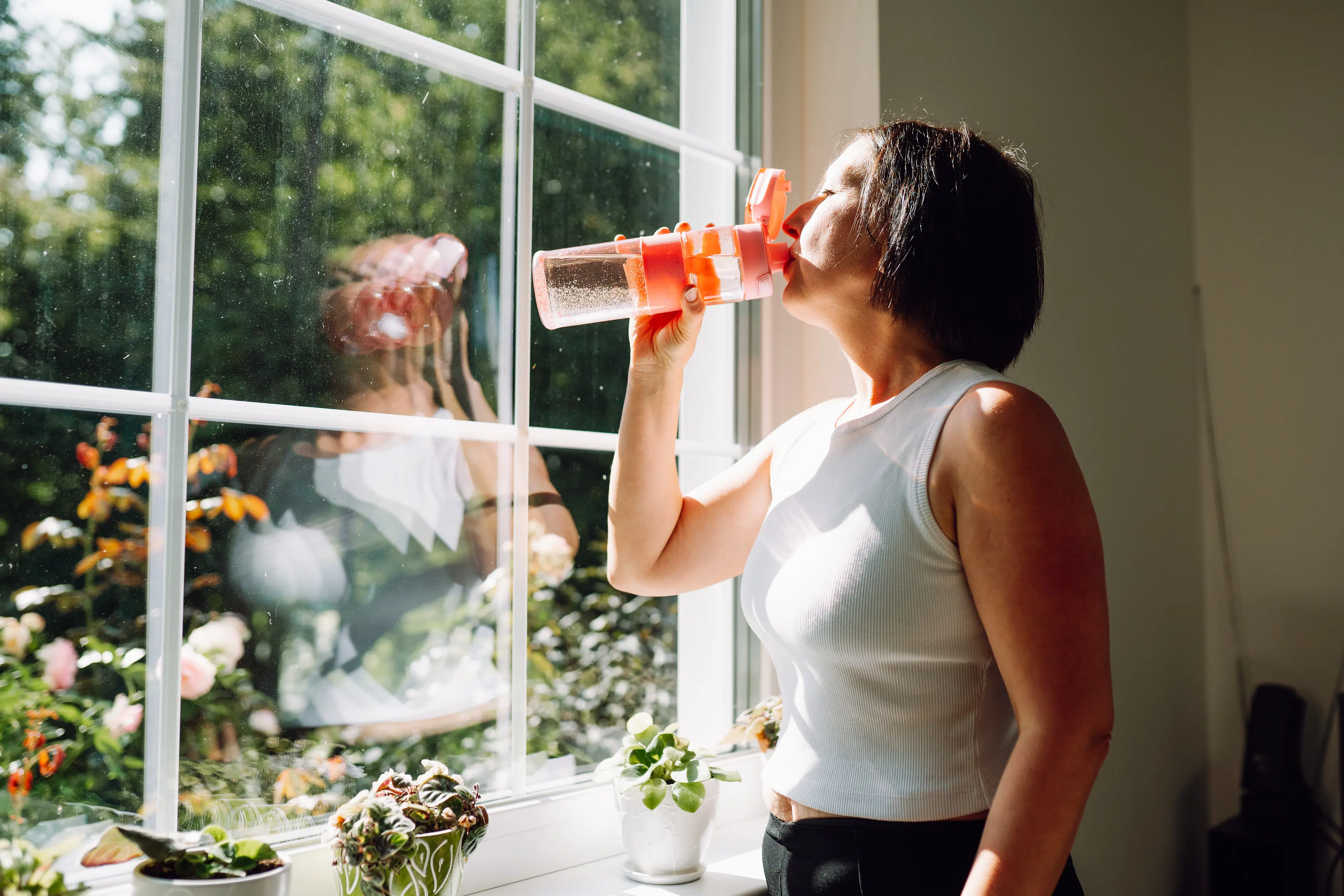A woman staying hydrated by drinking out of a clear reusable water bottle at home.