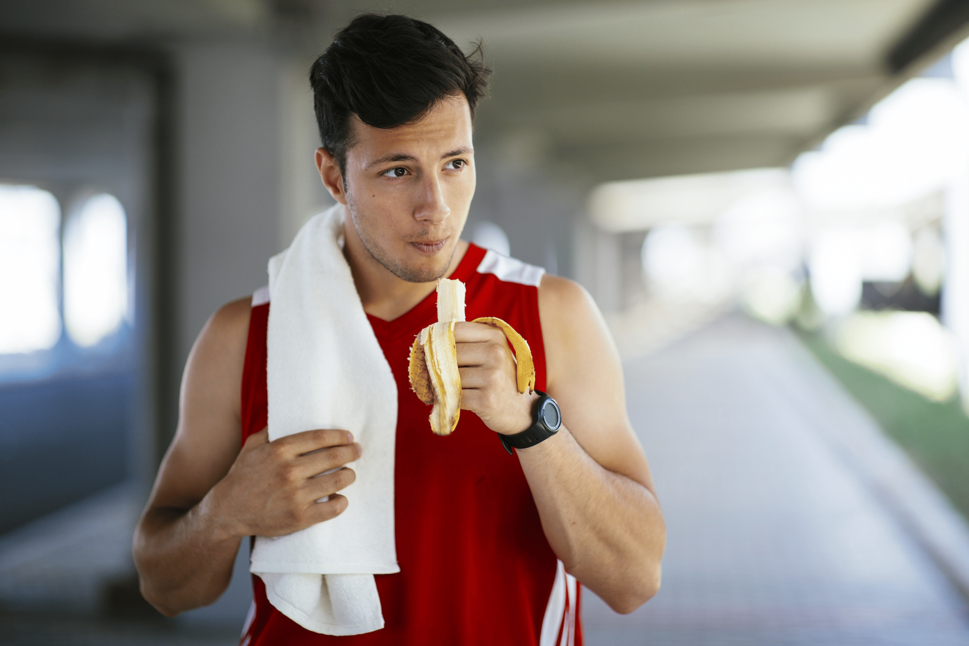 An athlete eating a banana after a workout while walking outside. He is wearing a red athletic tank top and has a white sweat towel over his shoulder.