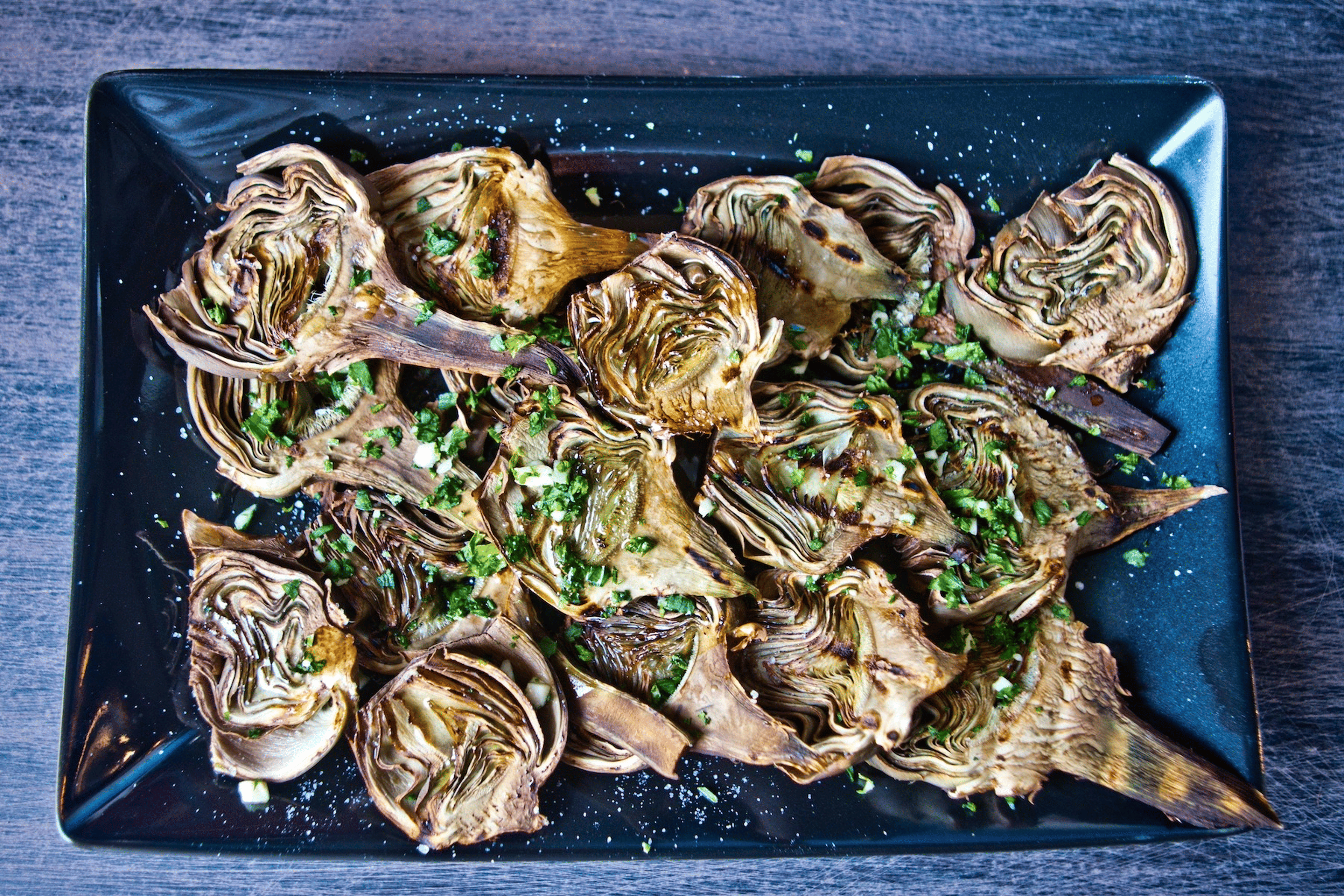 Flat-lay shot of baked artichokes on a black serving platter with herbs. Artichokes are a high-protein vegetable.