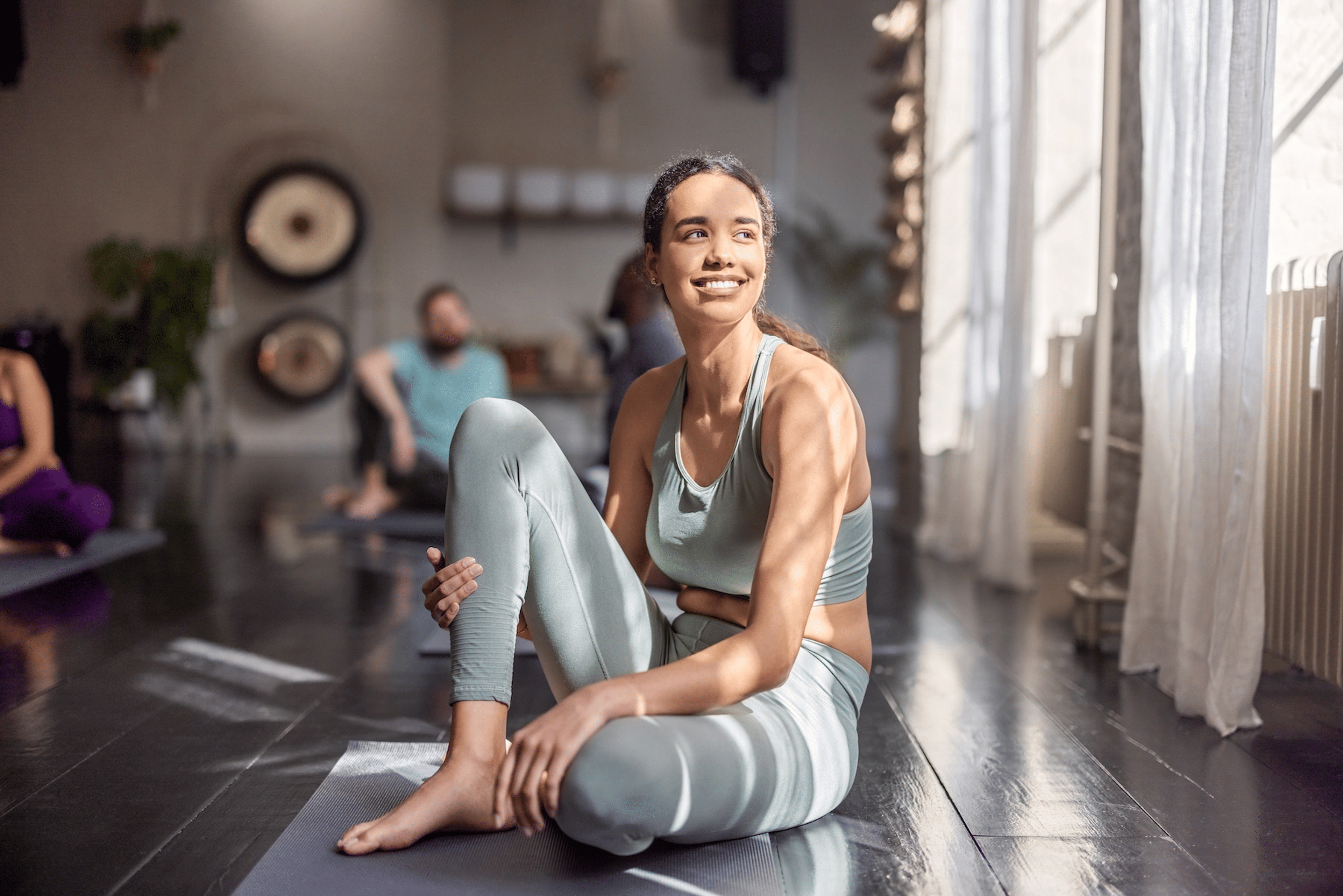 A woman sitting down on a yoga mat after exercising, smiling and looking away.