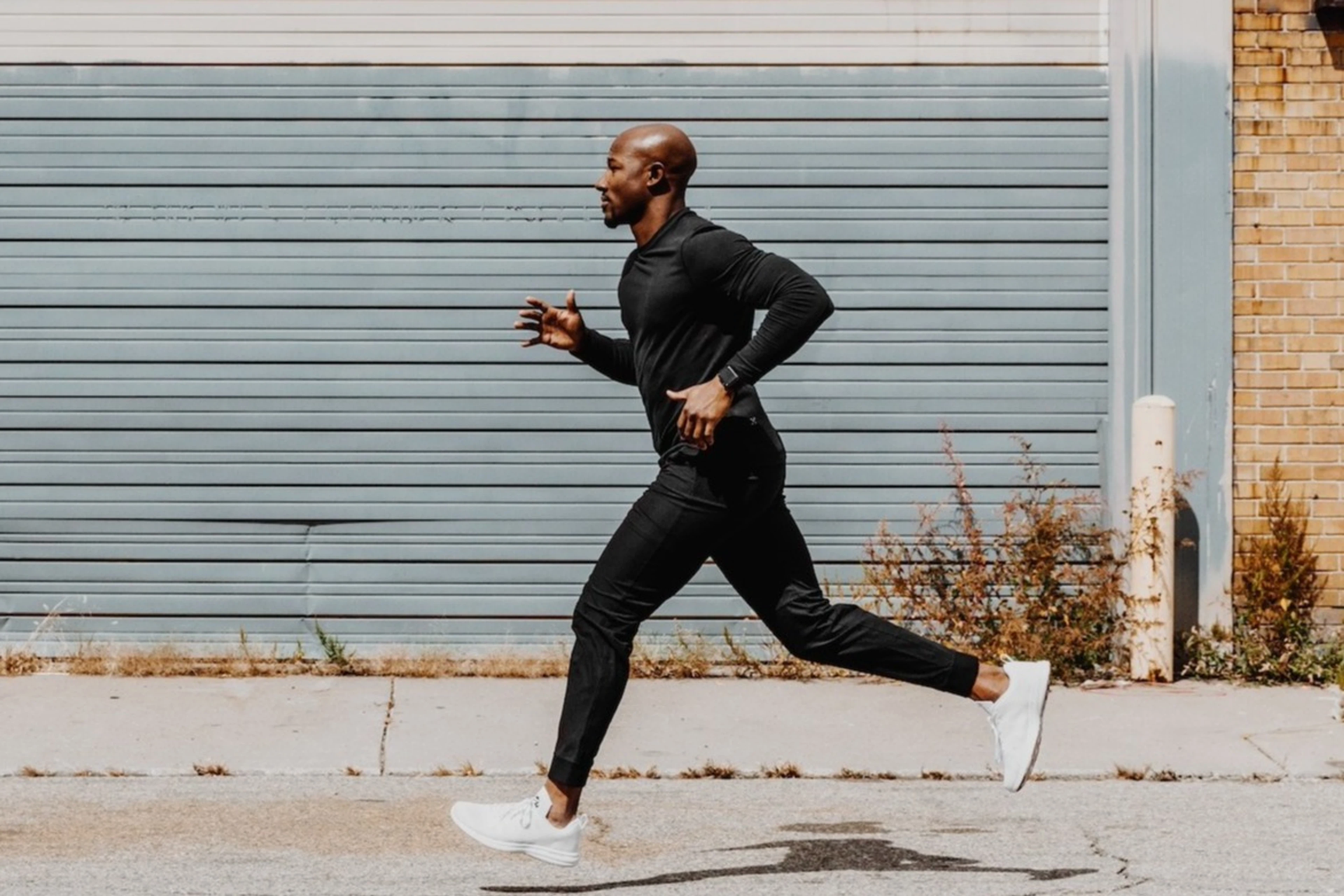 A man on a long run outside. He's wearing all black and jogging in front of an industrial building.