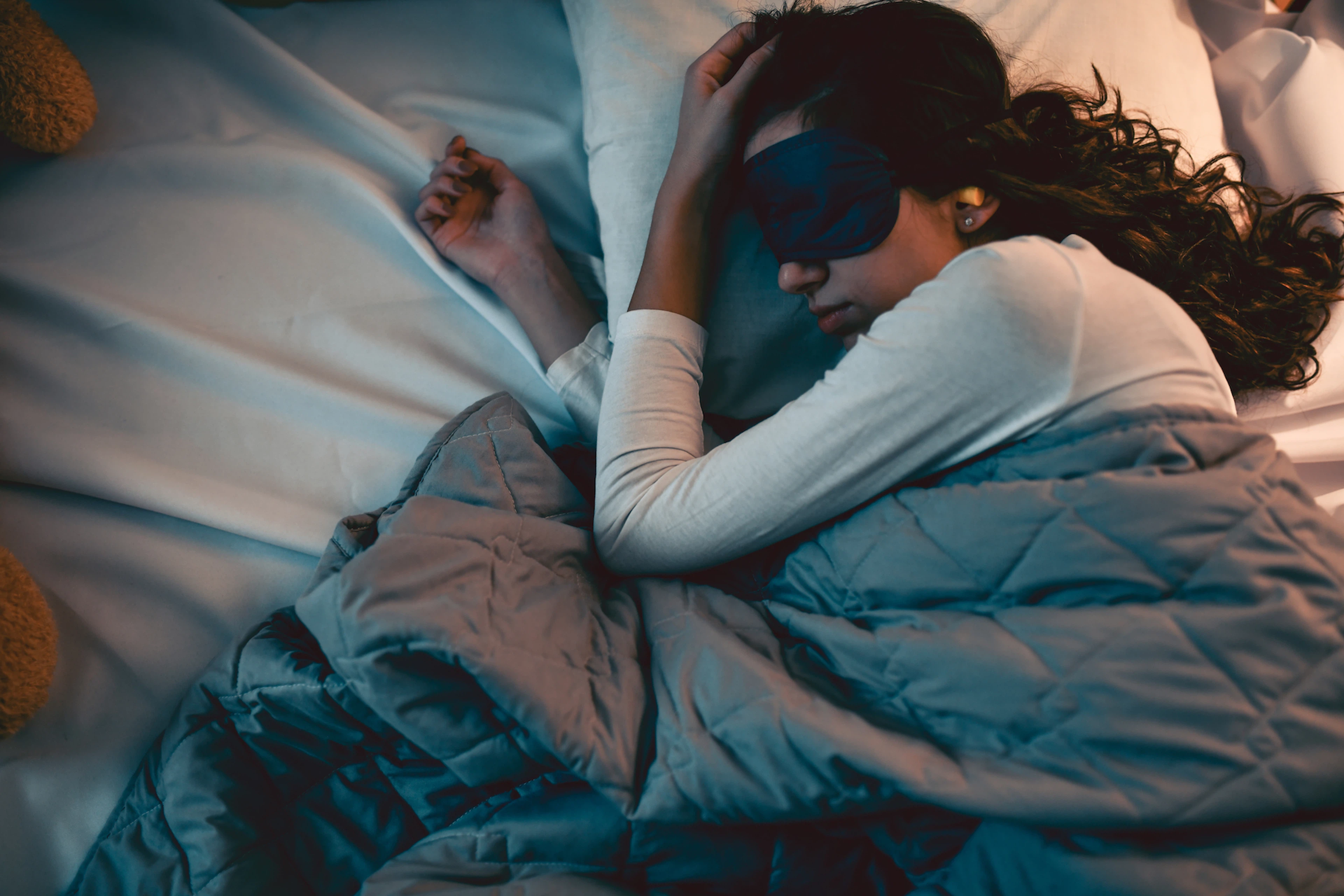 A woman sleeping in bed peacefully while wearing an eye mask and snoozing under a weighted blanket. Weighted blankets offer many benefits.