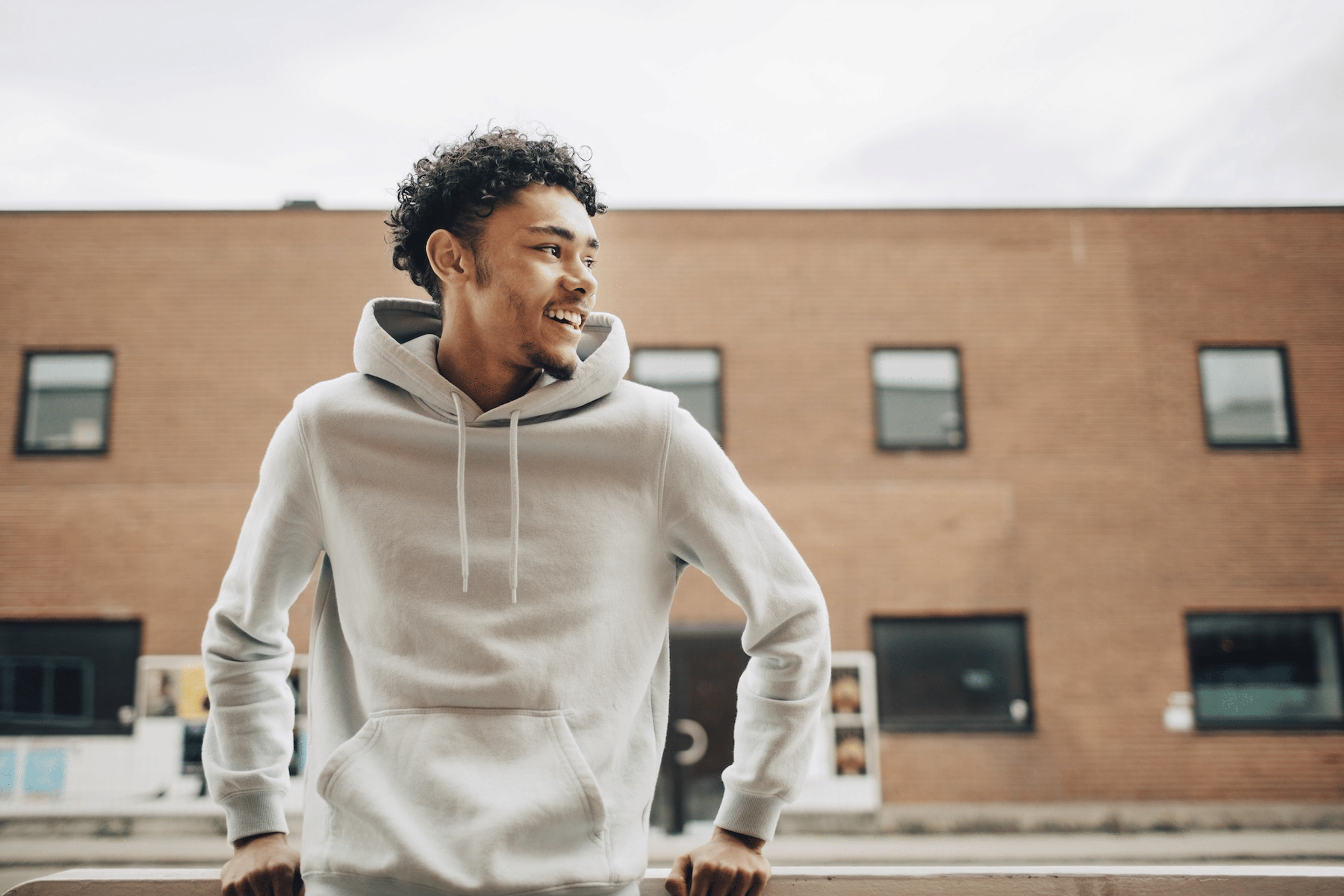 A man in a hoodie smiling and leaning against a railing outside. He's looking to his left.