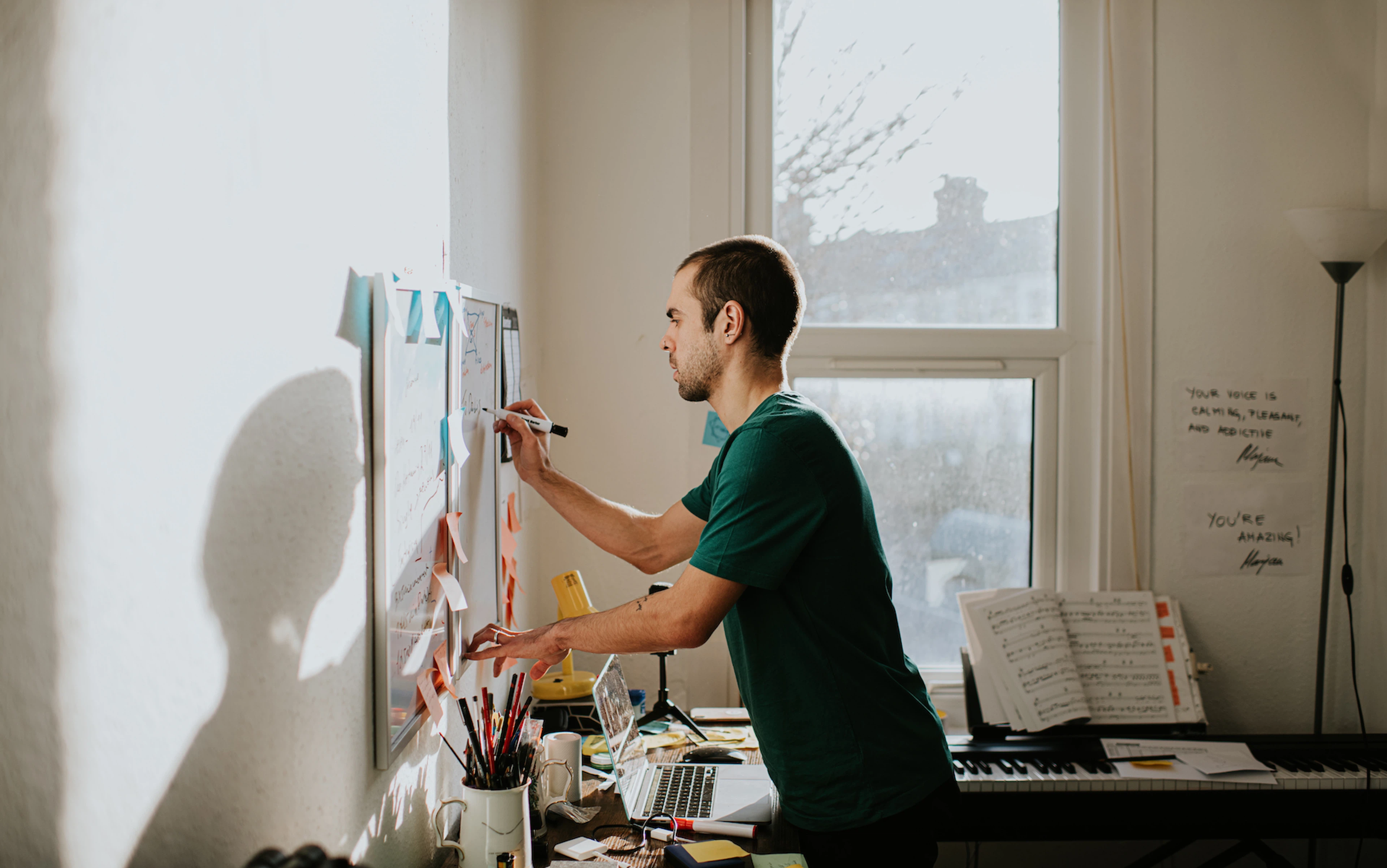 A man leaning over his desk and writing on a whiteboard in a sunny home office room. Learn "how long does it take to form a habit?" in this article.