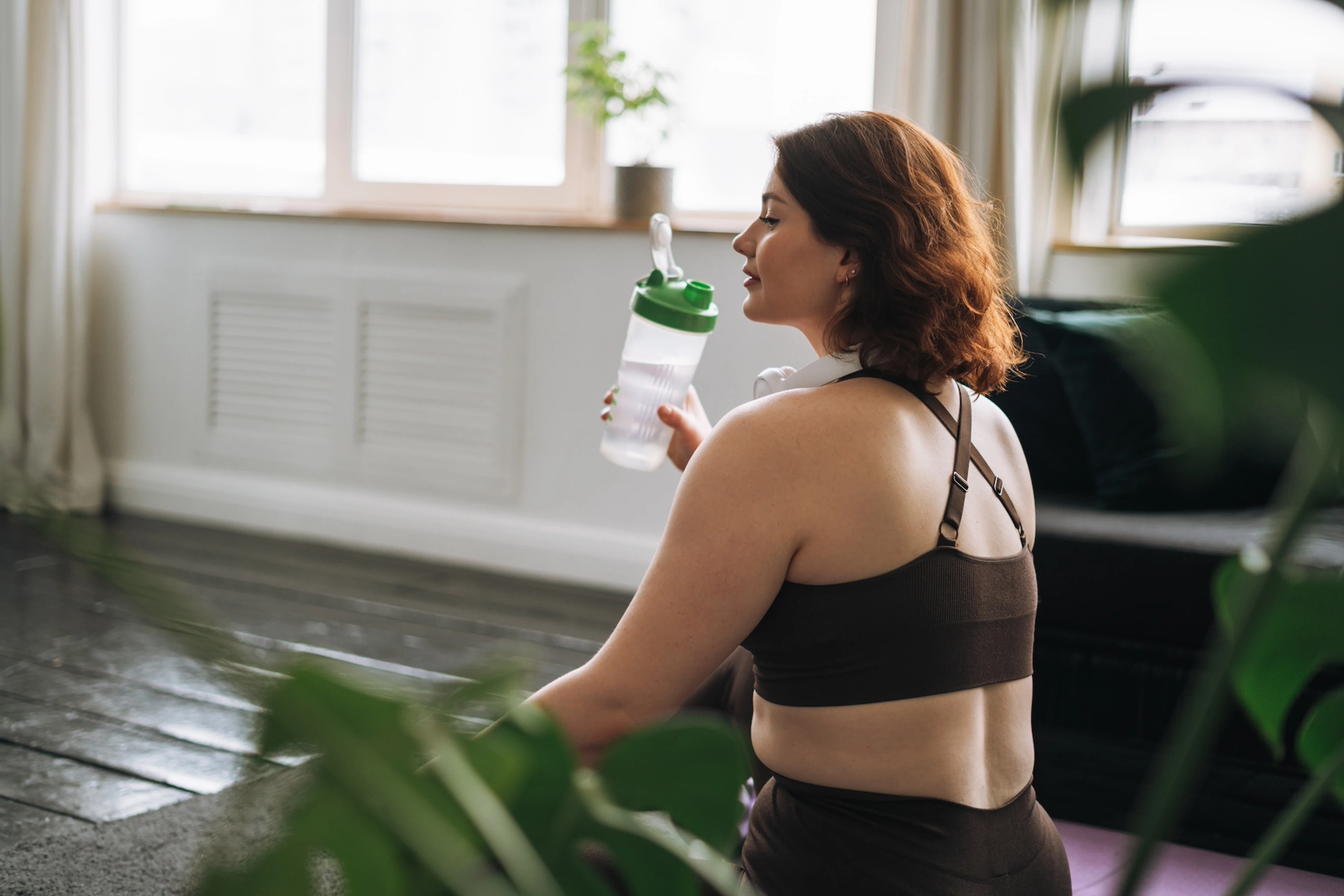 A woman drinking water out of a shaker bottle. She's wearing activewear and sitting at home.
